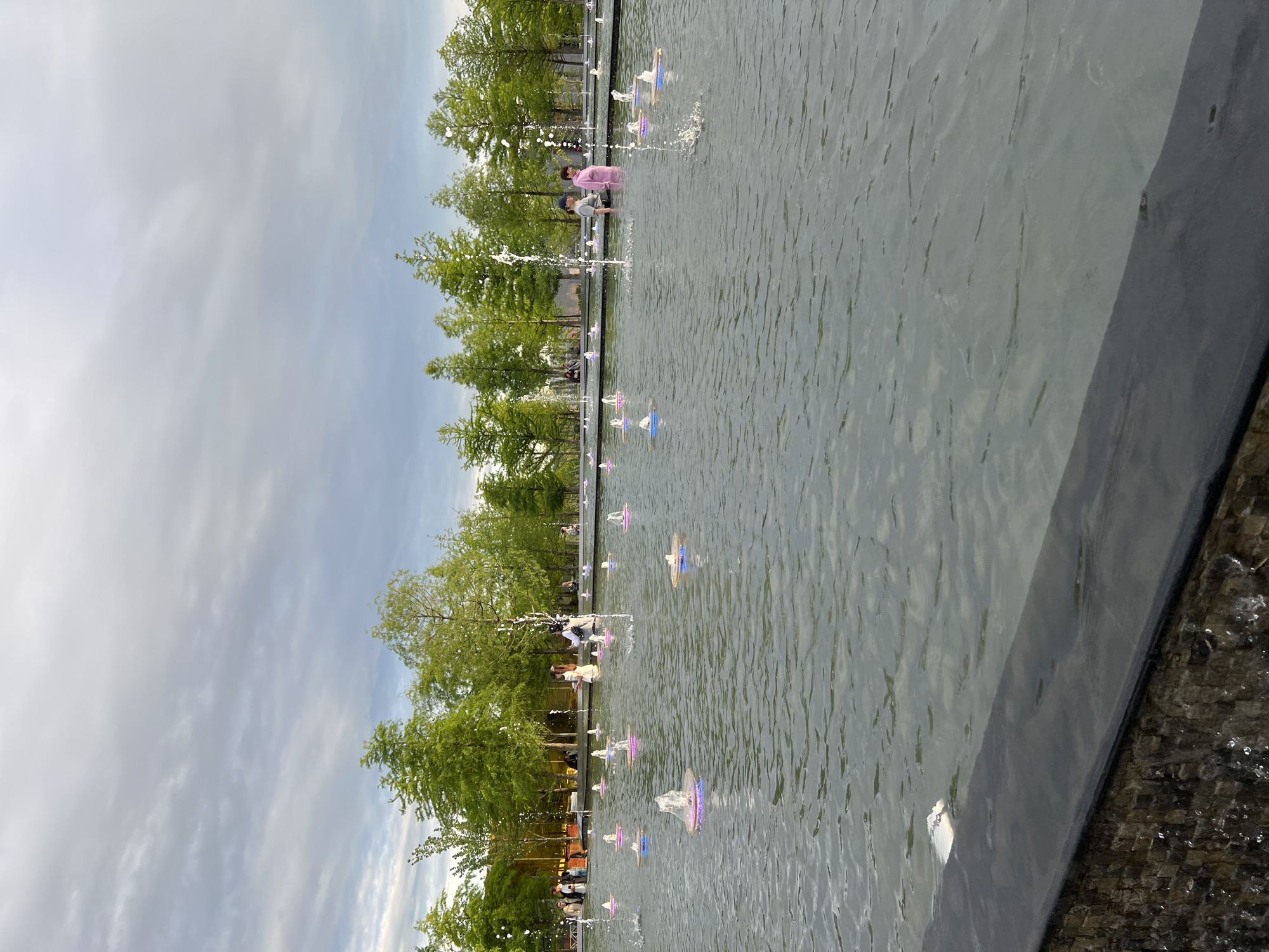 The image depicts a water fountain park in Krasnodar, Russia. Trees line the left side of the fountain, leading to a slightly overcast sky. People are walking in and around the fountain. The fountain itself has water jets and illuminated lights. The right side of the image shows a concrete edge of the fountain structure.