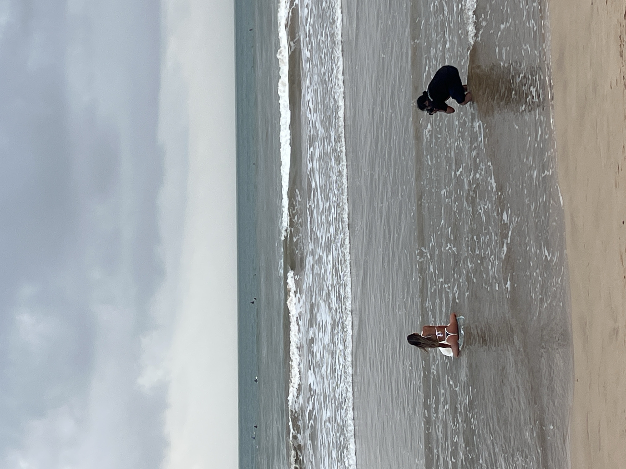 A photographer is kneeling in the shallow water of a beach in Peleana, Sri Lanka, taking a picture of a woman sitting on a surfboard in the ocean. The woman is wearing a white bikini. The sky is overcast, and there are waves breaking in the ocean. Several surfers are visible in the distance. The sand in the foreground is wet from the tide.