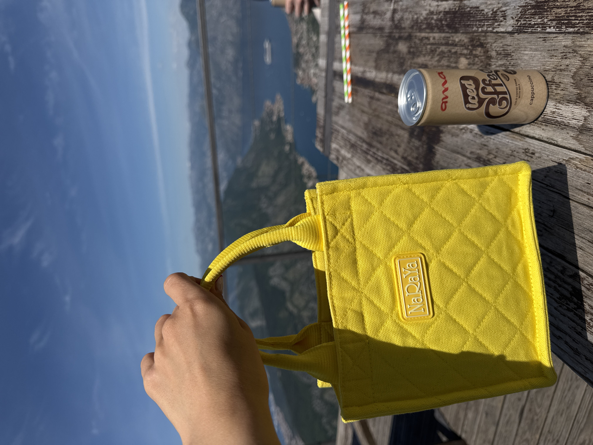 An outdoor scene captured at Žanjev Do, Montenegro features a yellow handbag held by a person's left hand, placed on a wooden surface. Next to the bag, there is a can of "ama Iced Coffee Cappuccino" and two drinking straws with red, green, and white stripes. The background reveals a mountainous landscape with a body of water between the mountains, under a clear blue sky. The location and perspective suggest a viewpoint from a height, overlooking the Žanjev Do region.