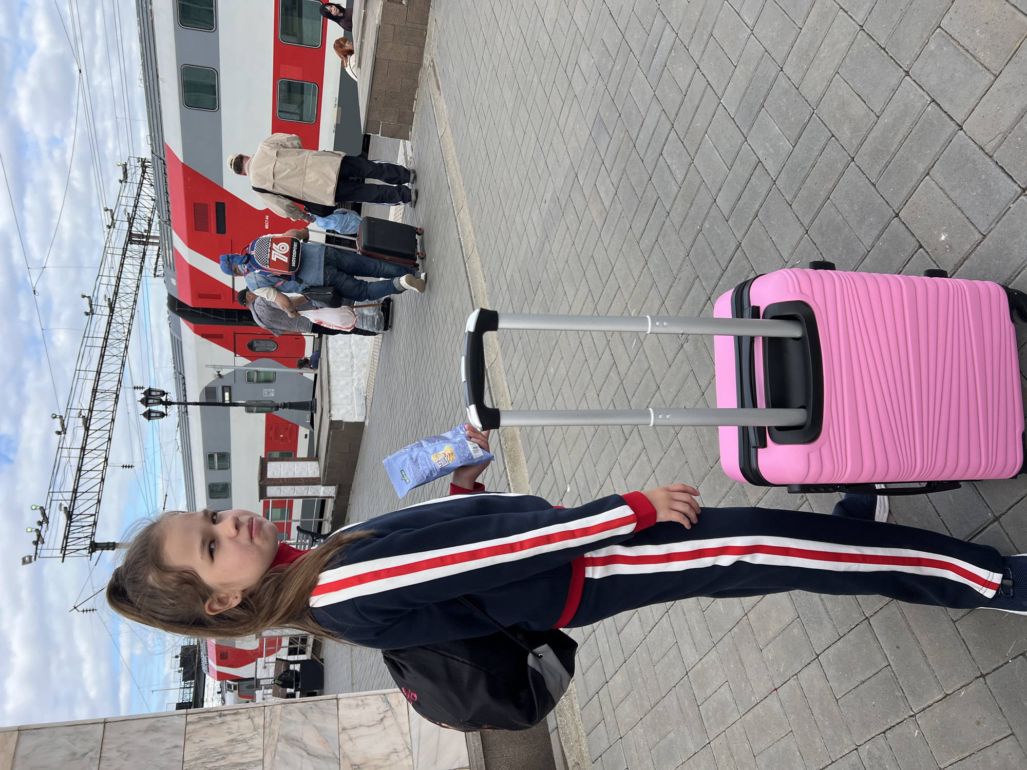 In Izhevsk, Russia, a young girl with her hair in a high ponytail stands on a paved train platform next to a pink suitcase. She is wearing a dark blue tracksuit with red and white stripes down the sleeves and pant legs, and a red turtleneck shirt. She holds a bag of snacks in her right hand. Behind her, a red and white train is stopped at the station, with several people boarding or disembarking. The sky is partly cloudy.