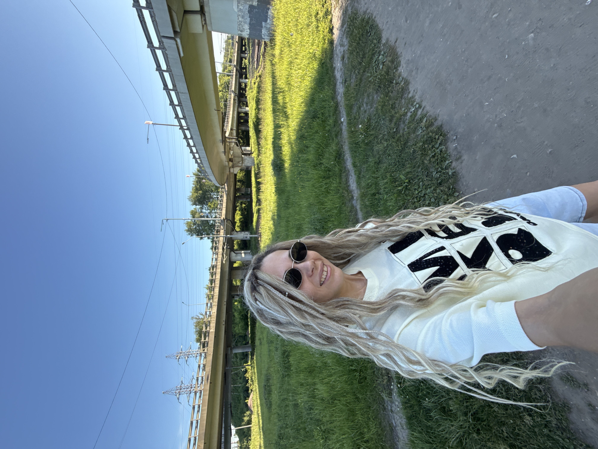 A female subject, estimated to be in her late 20s to early 30s, is captured in a selfie-style composition outdoors in Rusanovo, Russia. She has long, wavy blonde hair interspersed with lighter-toned dreadlocks or braids. Her eyes are obscured by dark, oval-shaped sunglasses, and she exhibits a smile, revealing her teeth. She wears a light-colored, long-sleeved top featuring a prominent black graphic design, which appears to include text such as "MIKE" and potentially sequins. A portion of light-colored lower garments is visible. Her left arm is extended towards the camera, indicating she is holding the device.
The background consists of a vibrant green grassy area, illuminated by direct sunlight that casts distinct shadows, suggesting either morning or late afternoon. Multiple concrete overpasses or bridges with railings are prominent in the mid-ground and background, supported by concrete pillars. Overhead power lines traverse the clear blue sky, originating from utility poles and two distinct metal power transmission towers located in the far left. Streetlights are mounted on poles along the bridge structures. A dirt path runs through the immediate foreground.