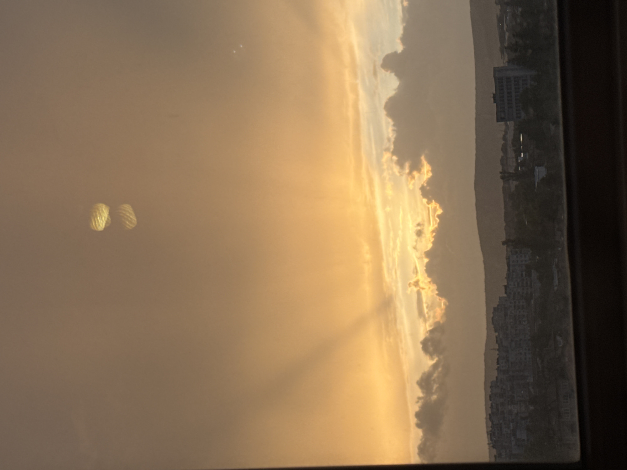 The image depicts an urban landscape at sunset viewed through a window. The sky is dominated by orange and yellow hues, with clouds partially obscuring the sun. Buildings and structures are visible in the distance. There are no people depicted. The location is identified as Lovech, Bulgaria based on contextual knowledge. Two circular light reflections are visible on the window.