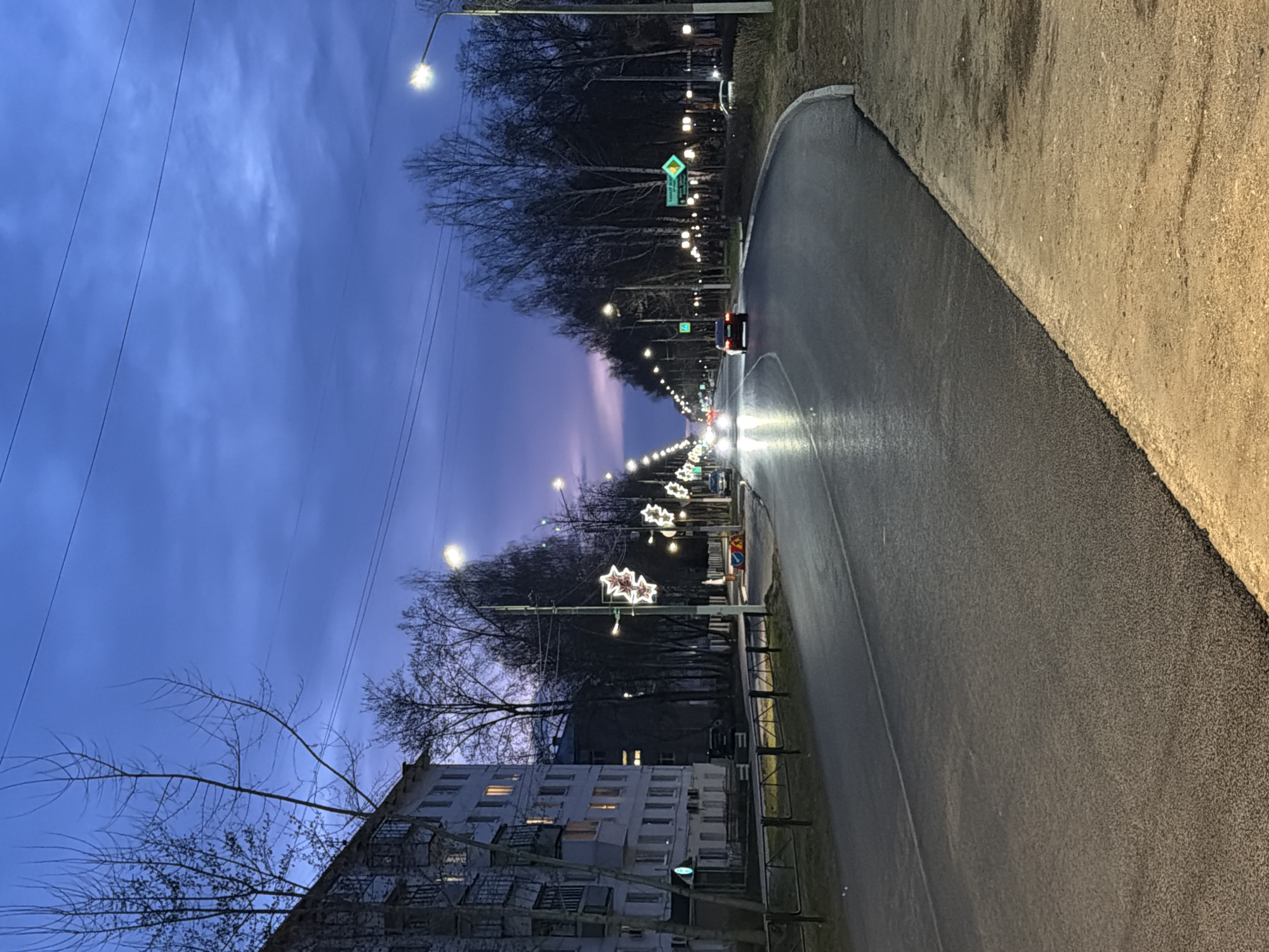 This nighttime photograph captures a serene urban street scene in Chaikovsky, Russia. The perspective is looking down a long, well-lit asphalt road that curves slightly to the right, bordered by a dirt or gravel shoulder. The road appears freshly paved in some sections. Along the left side of the street, a line of bare trees stands, illuminated by numerous streetlights and decorative star-shaped lights attached to poles, adding a festive touch. A vehicle, likely a car, is seen driving away in the distance, its bright headlights creating light streaks on the wet-looking road surface.
To the far left, a multi-story residential building with several illuminated windows is partially visible. The sky overhead is a dark, clear twilight blue, with faint cloud formations. Power lines are visible stretching across the upper portion of the sky. A green street sign with a yellow directional arrow pointing right is noticeable, displaying white Cyrillic text that reads "Ленина ул." (Lenina Street). The overall impression is one of a quiet, illuminated street during the early evening hours.
