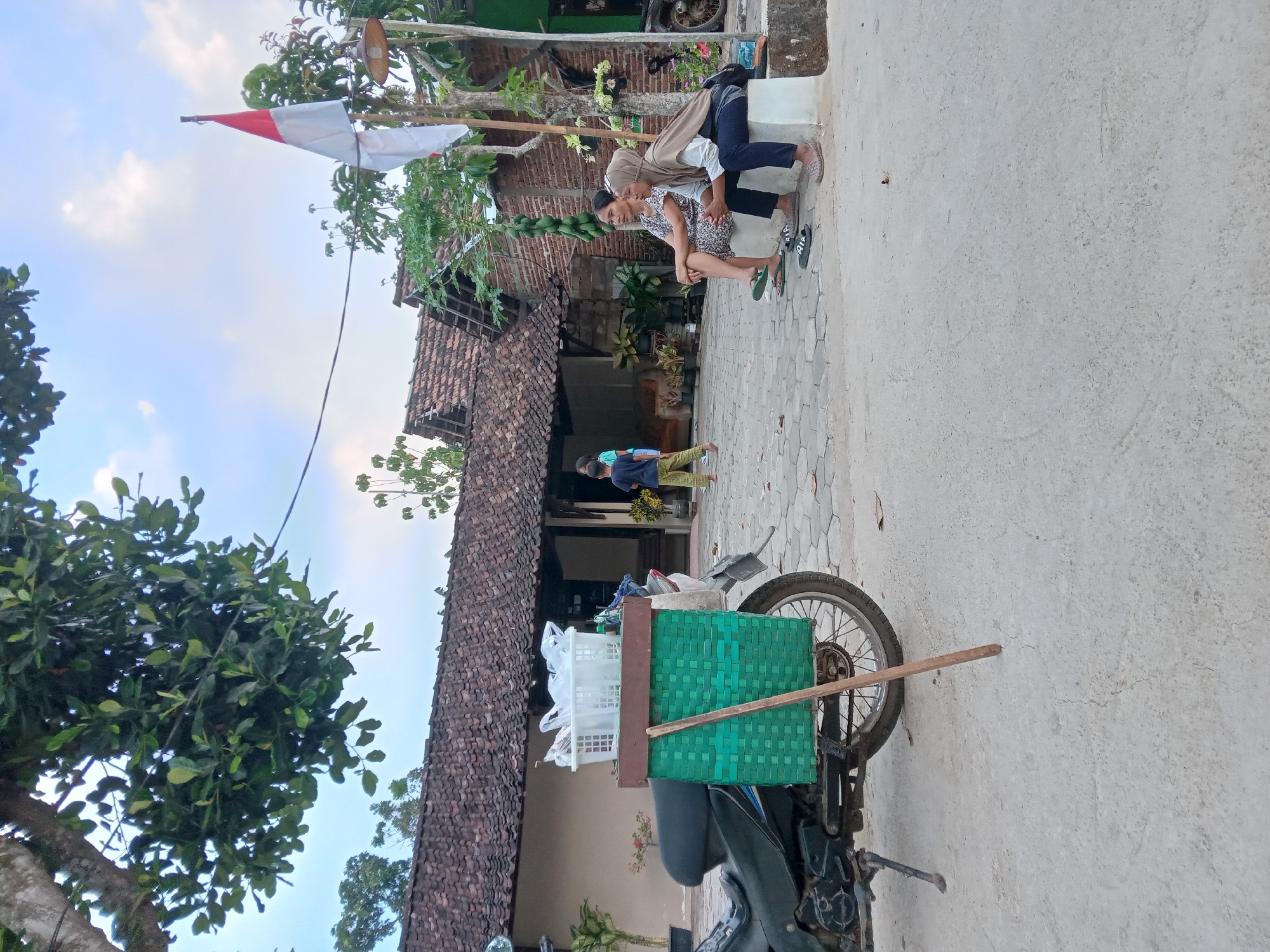 The image depicts a street scene in Kedunggudel, Indonesia, with a focus on a motorcycle parked on a paved surface. The motorcycle is equipped with a large, green woven basket attached to its rear, containing a white plastic basket filled with items. Behind the motorcycle, two women are seated on a low concrete structure. One woman wears a patterned dress and the other a hijab and a long-sleeved shirt. They are looking towards the right of the frame. Further back, a house with a tiled roof and brick walls is visible. A flagpole with the Indonesian flag is planted near the house. In the background, several people, including children, are walking and standing. The sky is partially cloudy, and the overall lighting suggests it is either early morning or late afternoon.