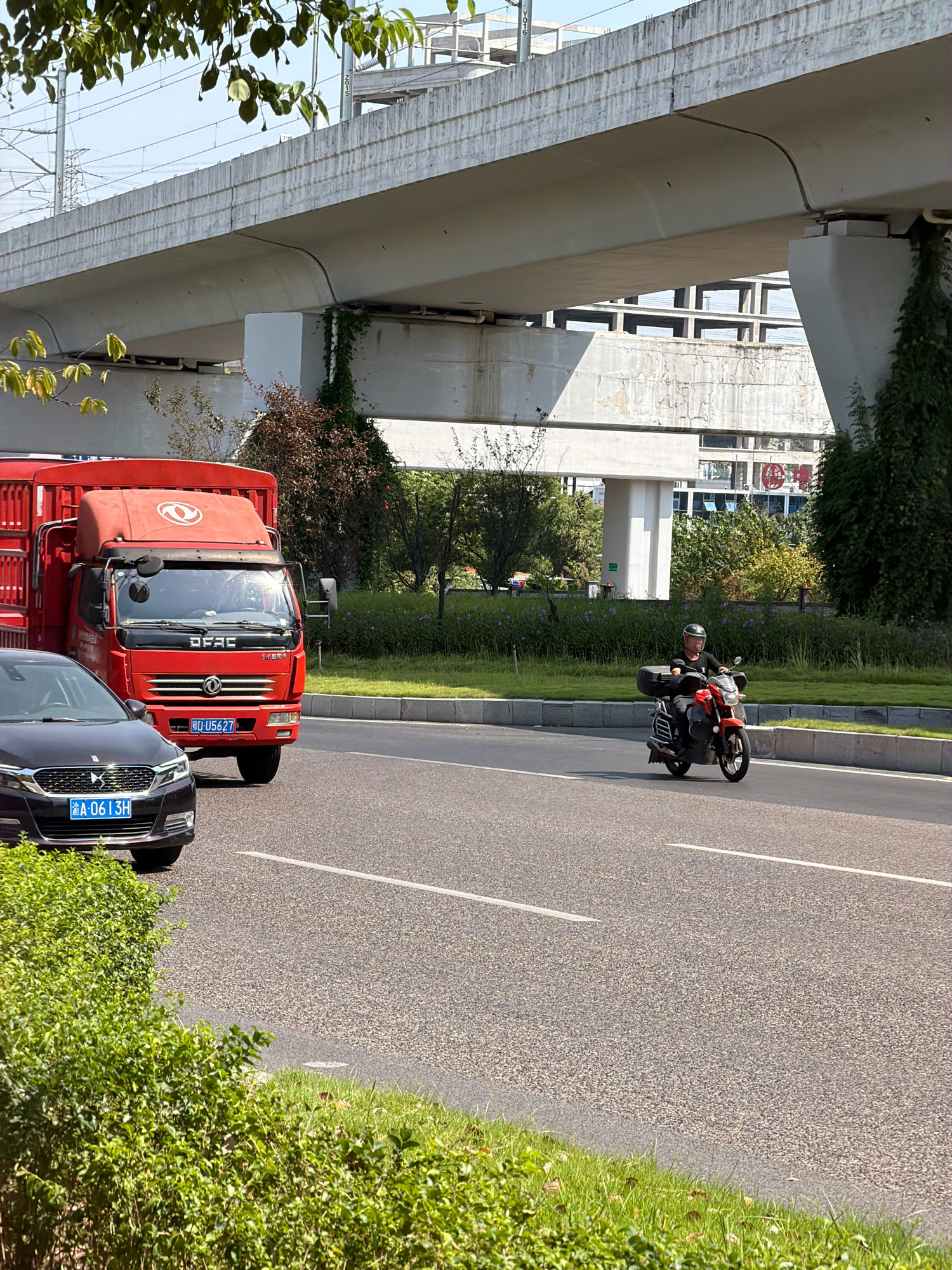 The image depicts a street scene in Jiangjin, China, under an elevated overpass. In the foreground, a dark colored car is parked on the left side of the road, displaying a license plate that reads "A-0613H". To its right, a red truck with a logo on its hood and a license plate reading "D-U5627" is traveling on the road. Further ahead, a person wearing a helmet and a black shirt is riding a red motorcycle. The environment includes a green grassy median separating the lanes of traffic, with trees and bushes visible. The overpass, made of concrete, runs across the upper portion of the image, with visible electrical wires and support structures. Buildings and more greenery are seen in the background. The sky is clear and blue.