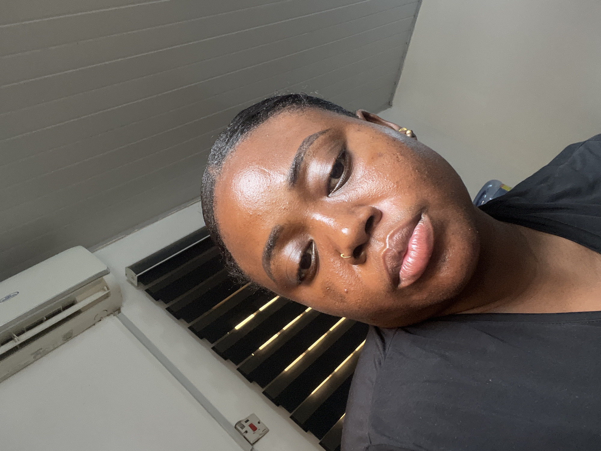 A close-up, angled shot of a woman with dark skin and short, dark hair. She is wearing a black shirt and a gold nose ring in her left nostril, as well as gold earring. Her head is tilted to the right. The background includes a white ceiling with horizontal ridges, an air conditioning unit, and a window with vertical blinds. The image appears to be taken indoors. There are no other people visible. The location cannot be determined to be in Abuja, Nigeria, based on the image alone.