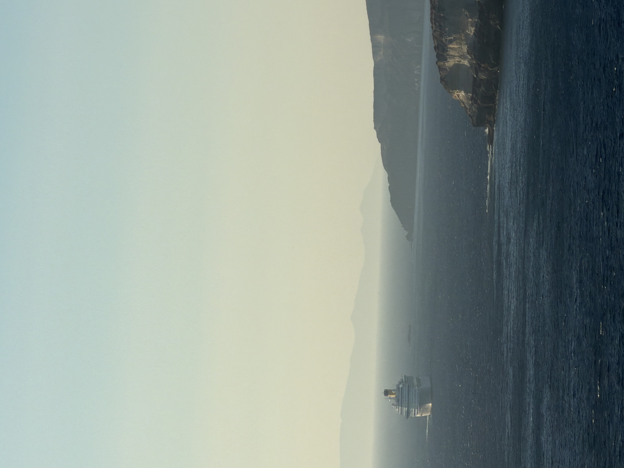 The image shows a cruise ship sailing in the Aegean Sea, approaching the coast of Santorini. The cruise ship is white with a yellow funnel and is positioned in the left-center of the frame. The sea is a deep blue with gentle waves. In the background, a series of hazy, layered mountains are visible, suggesting a coastal landscape. The sky is a pale, gradient blue, transitioning to a light yellow near the horizon, indicative of a late afternoon or early morning light. On the right side of the image, a rocky cliff face juts out into the sea, with a lighter colored rock formation at its edge. The scene is serene and expansive, conveying a sense of travel and natural beauty. The location is the sea off the coast of Thira Municipal Unit, Greece, likely near the caldera of Santorini.
