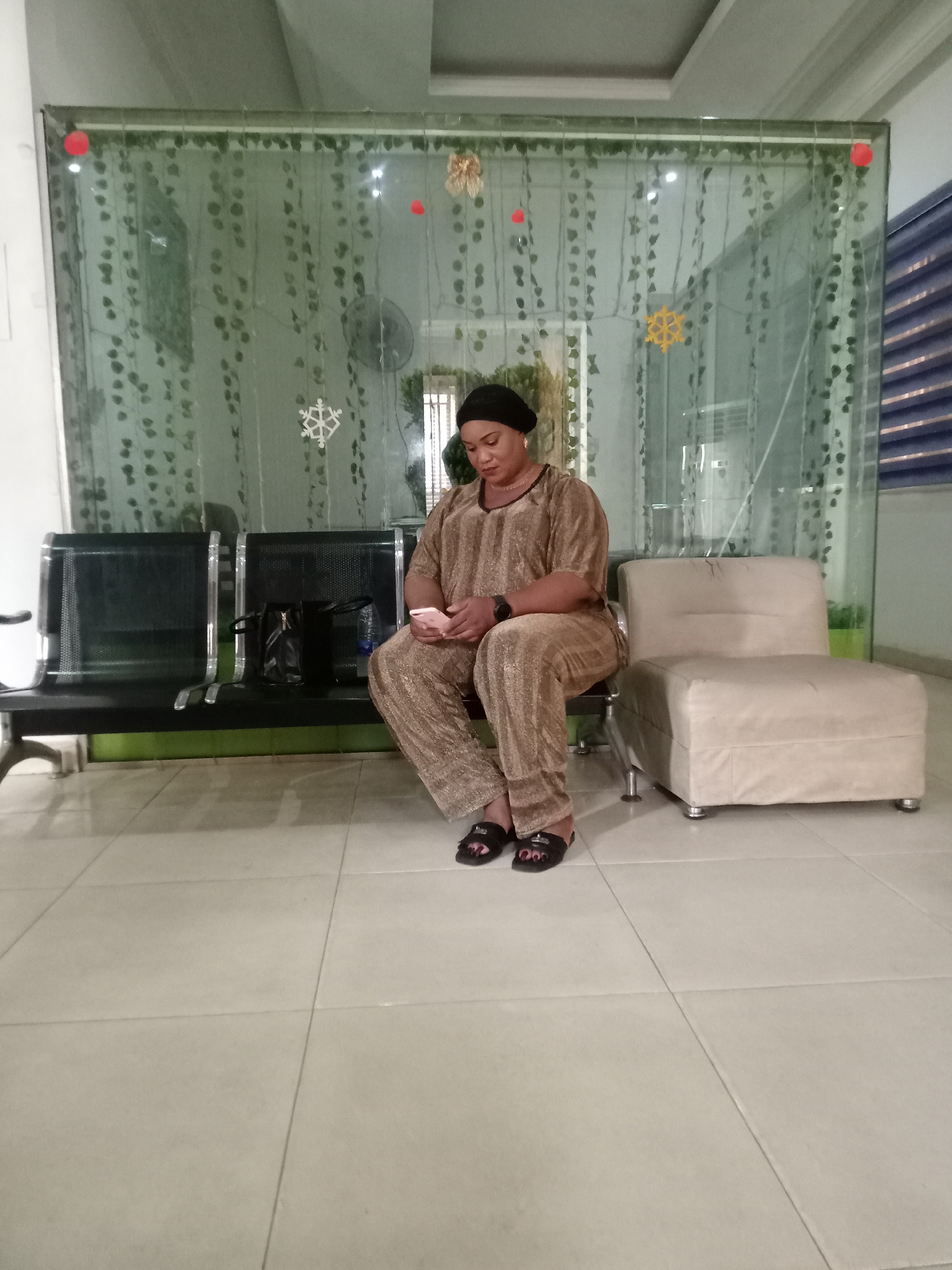 A woman in a matching brown top and pants sits on a black metal bench in what appears to be a waiting area. She is looking at a mobile phone in her hands. Next to the bench is a black handbag and a tan, single-seat cushioned chair. Behind her is a decorative glass partition adorned with faux ivy and holiday ornaments. The floor is tiled. The setting is Gwarinpa, Nigeria.