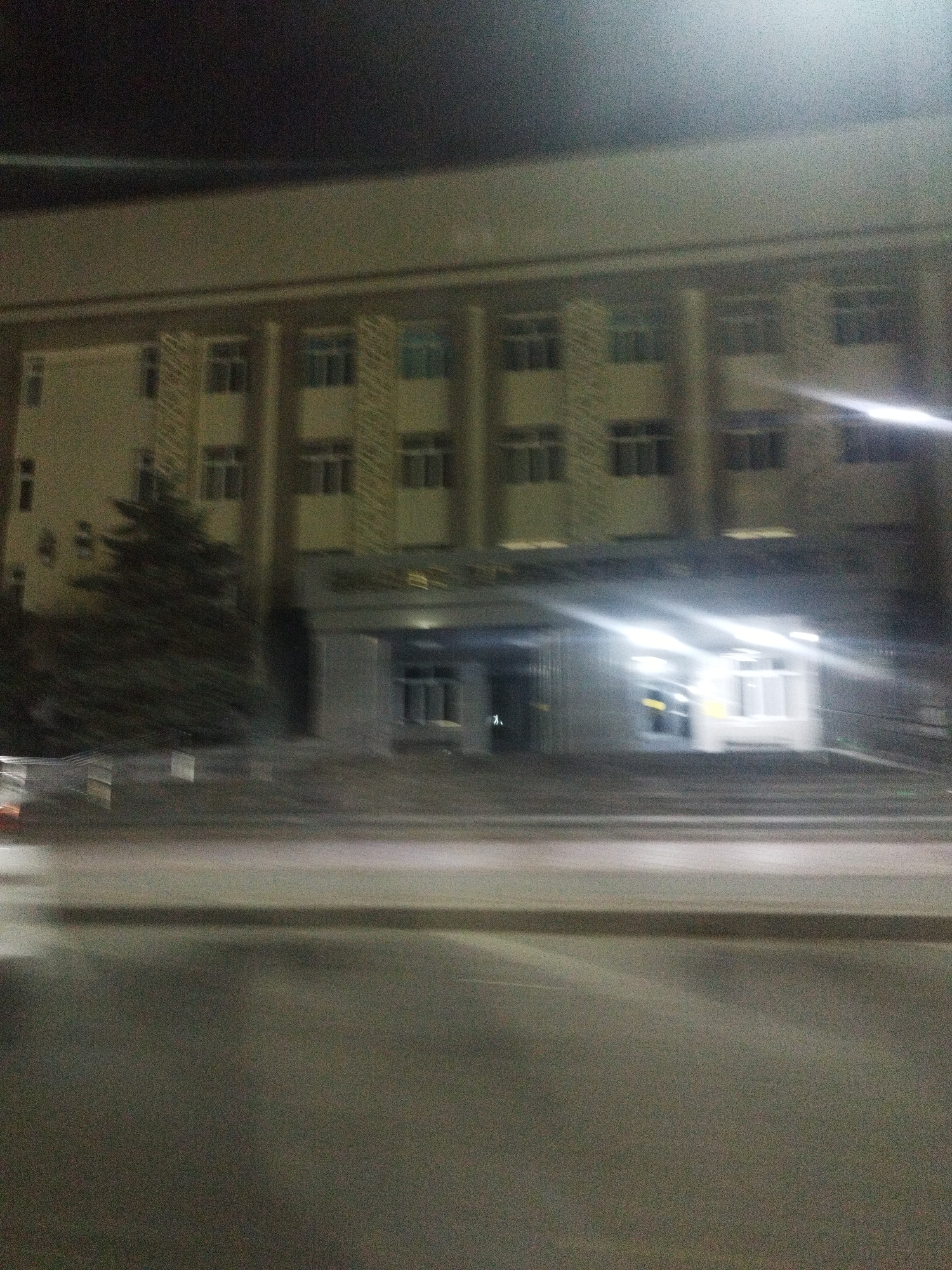 The image shows a building with multiple stories at night. The building has a beige facade with a grid of windows on each level. There are some trees in front of the building. The foreground shows a street with some blur due to motion. The image appears to be taken from a moving vehicle. Based on the information provided that the location is in the city of Shymkent, Kazakhstan, it is likely a government building or an educational institution. The lighting suggests an urban setting.