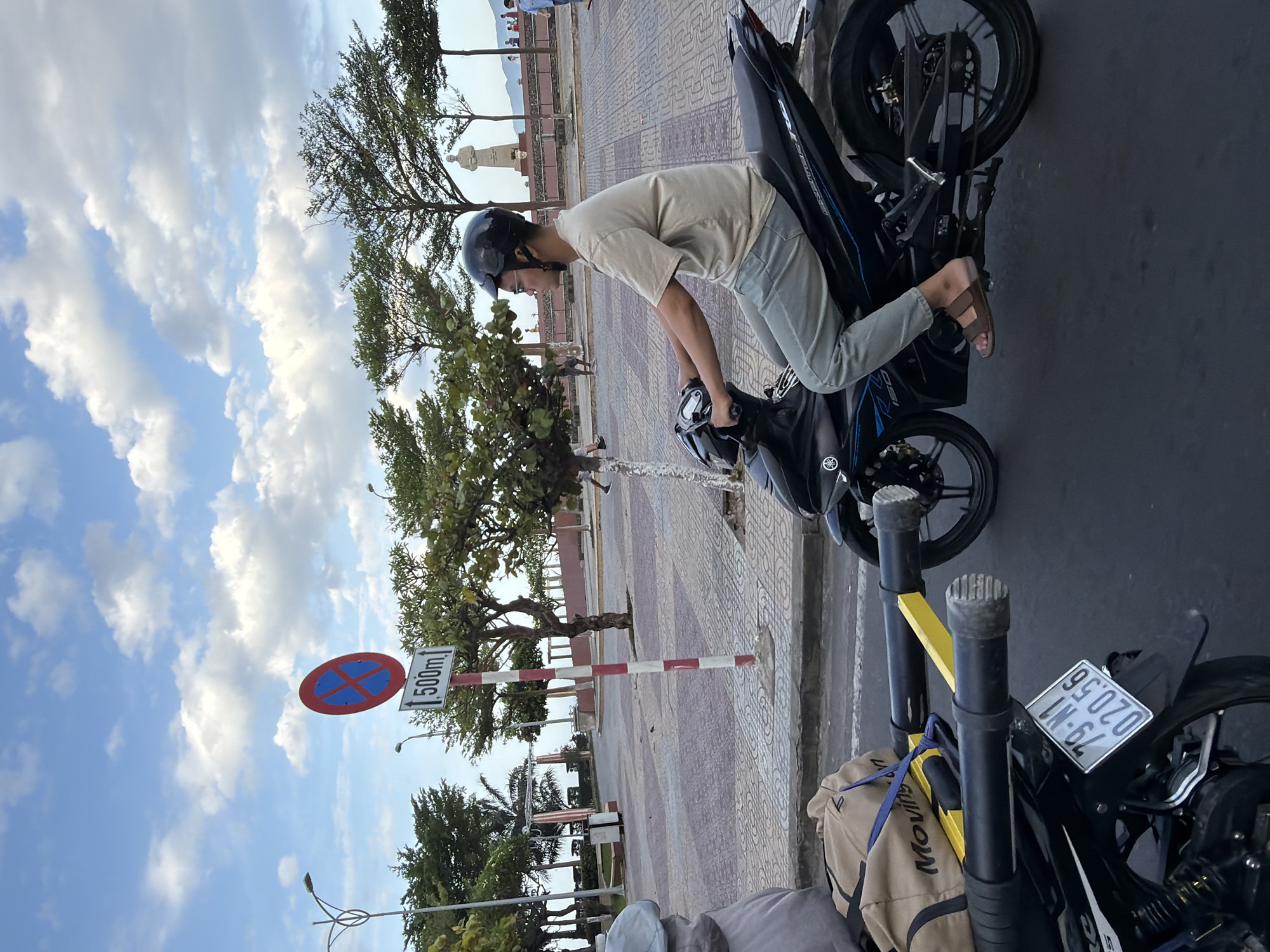 In Nha Trang, Vietnam, a person wearing a black helmet and glasses rides a black motorcycle. They are dressed in a light brown t-shirt and light gray pants, with brown sandals on their feet. The motorcycle has a license plate reading "79-N1 020.56". In the foreground, a beige bag with "Moving" printed on it is strapped to the motorcycle. Behind the rider, a red and white striped pole supports a traffic sign with a red circle and a blue cross inside, indicating no parking, and below it, a white sign with "500m" and an upward arrow. The background features a paved area with a checkered pattern, trees, streetlights, and a statue of a man. In the distance, more trees and buildings are visible, along with a cloudy sky.