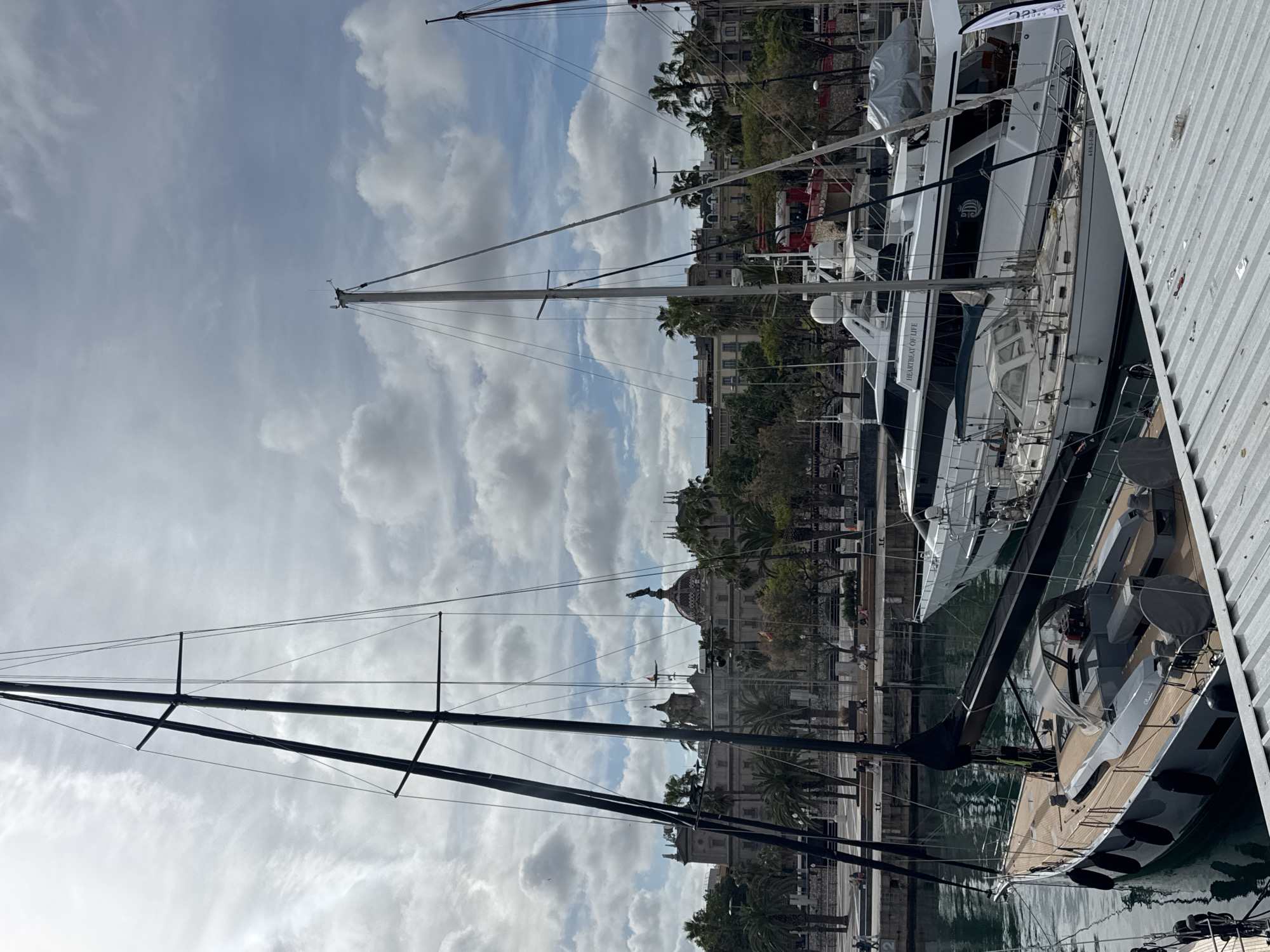 This outdoor image depicts a marina in Barcelona, Spain. The viewpoint is from a dock, looking towards several docked yachts and boats. In the background, buildings are visible, interspersed with palm trees, adding a Mediterranean cityscape feel.
The sky is overcast with scattered clouds. Two prominent masts of sailing yachts extend upwards, reaching towards the cloudy sky. The overall color palette is dominated by whites, grays, blues, and natural wooden tones of the yachts, reflecting the sky and marina surroundings.