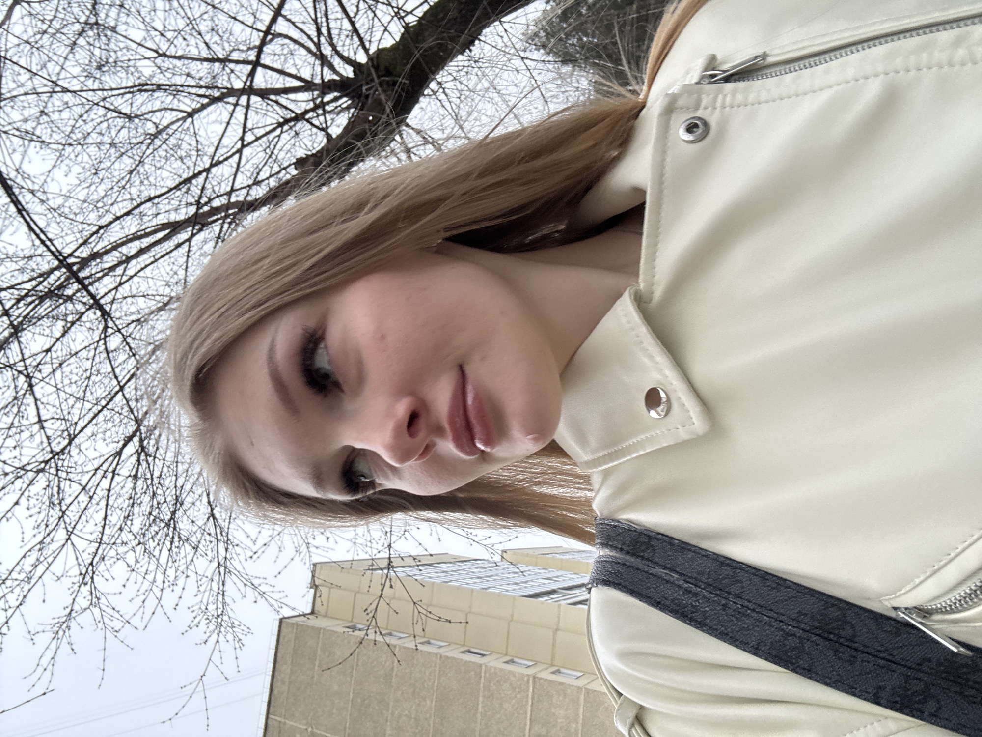 A woman is taking a selfie while outside. She is wearing a light-colored leather jacket and a dark strap across her chest, likely from a bag. The background shows a building with many windows and a bare tree with visible branches. Based on the architecture and the general environment, it's plausible that the location is Perm, Russia. The woman is the primary subject, and the building and tree are background elements. The action is the woman taking a selfie. There's no visible interaction with others in the frame. The environment is urban, likely cold given the bare tree, and possibly overcast based on the lighting.