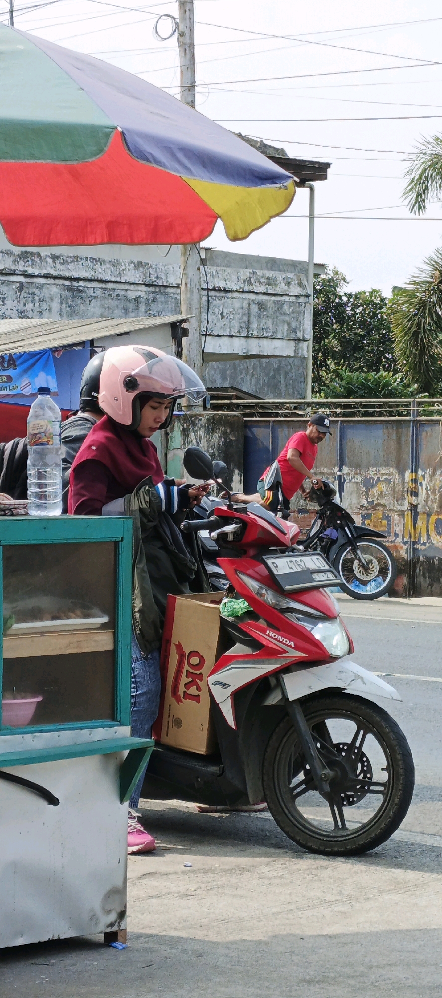 A street scene in Kasiyan, Indonesia, features a woman wearing a pink helmet and a maroon headscarf, sitting on a red and white Honda scooter. She is looking at her phone, which she holds in her hands. The scooter has a brown cardboard box with "OKI" printed on it strapped to the back. To her left, a street cart with a green frame displays food in clear containers. Behind her, another scooter is parked, and a man in a red shirt and a black cap is visible. A large, colorful umbrella, striped with red, yellow, blue, and green, is positioned above the scene. The background consists of a concrete wall and some vegetation.