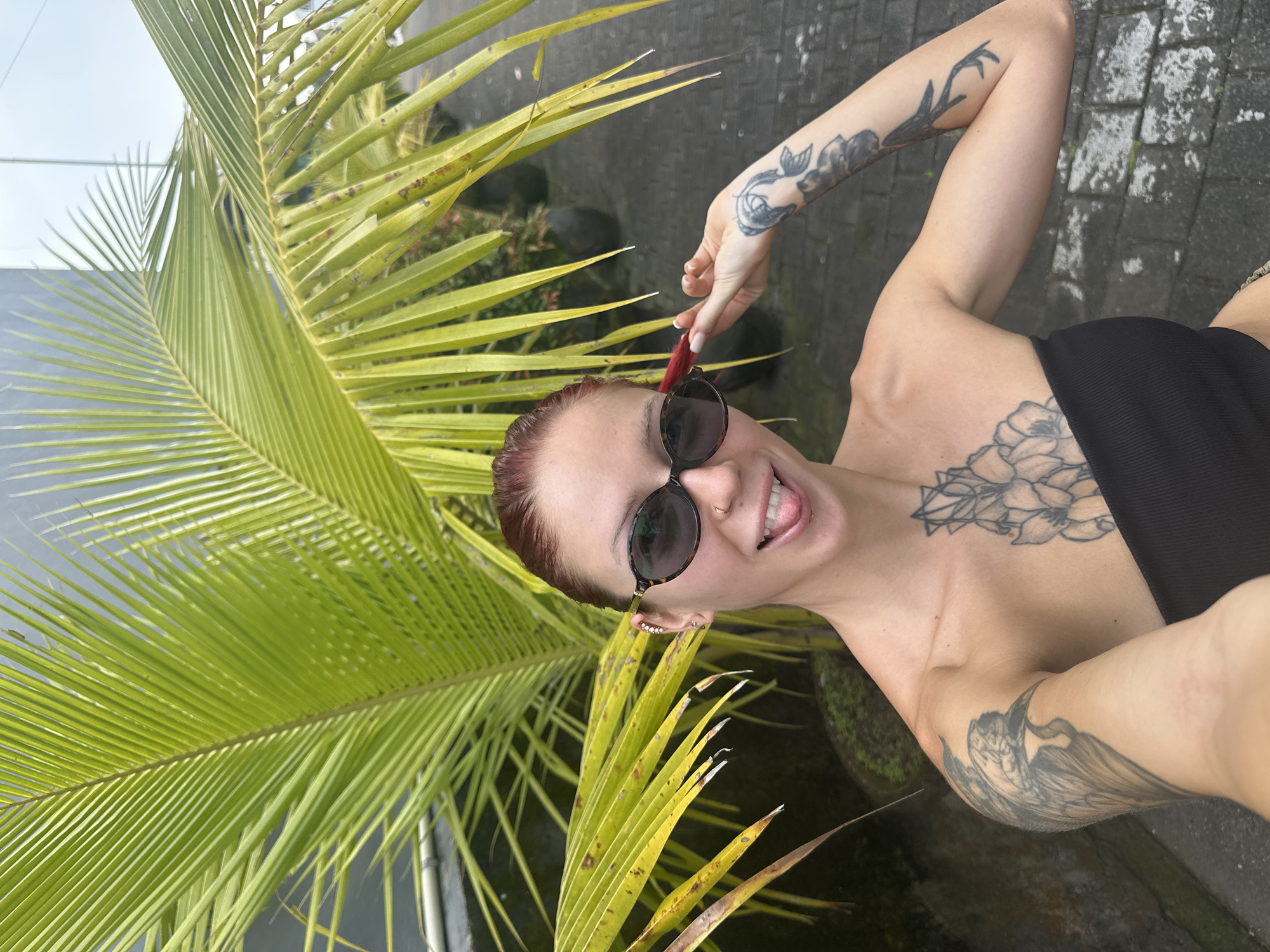 A female subject is depicted in a close-up, chest-up selfie format, standing outdoors in Dalung, Indonesia. She has auburn hair pulled back, wears dark oval sunglasses, and exhibits multiple tattoos: a large black-ink design of a bird on her upper left arm, a geometric floral pattern on her sternum/upper chest, and botanical/abstract designs on her right forearm and wrist. Ear piercings are visible, including a cuff of stones on her left ear and a small ring on her right nostril. She wears a black strapless top. Her expression is playful, with her tongue slightly extended, and her right hand holds a strand of her hair near her right temple. The background is dominated by large, vibrant green palm fronds, some displaying yellowish tips and minor brown discoloration. To the lower right, a paved surface composed of grey interlocking bricks or pavers is visible, alongside several dark potted plants lining what appears to be a path. A light grey building structure is partially visible in the upper left background.