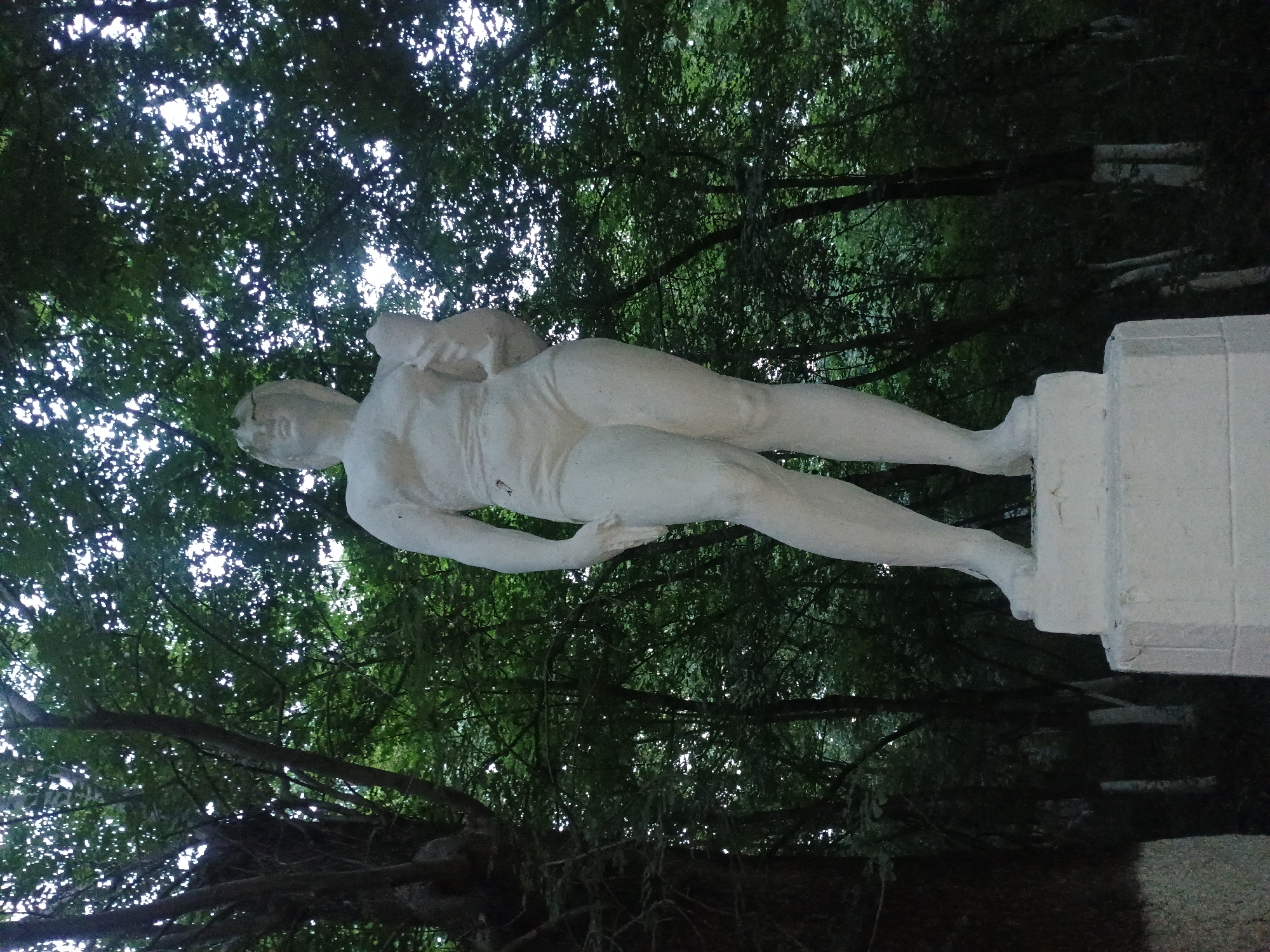 A full-body, white, plaster or concrete statue of a female figure stands on a white pedestal. The statue depicts a woman with short hair, a muscular physique, wearing shorts and a top that accentuates her abdominal muscles. She is holding an object in her right arm, possibly a vessel or a piece of fruit. The statue is positioned outdoors in a natural setting with dense green foliage and trees forming the background, suggesting it is located in a park or a wooded area. The lighting indicates it is daytime, with sunlight filtering through the leaves. The provided context indicates this statue is located in the city of Gusar, Azerbaijan.
