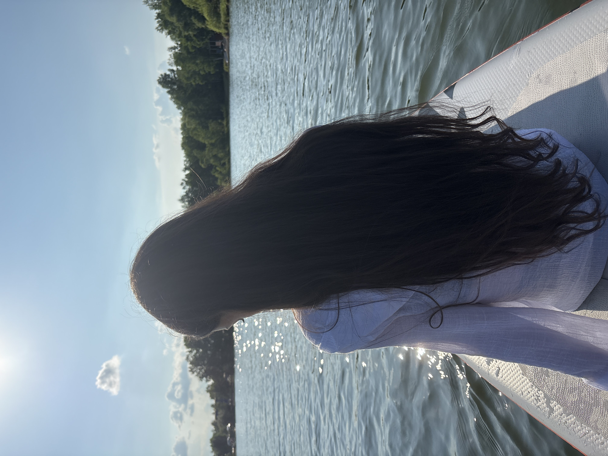A person with long, dark hair is sitting on a paddleboard on a calm lake, facing away from the camera. The person is wearing a white, loose-fitting shirt. The sun is shining brightly, creating a reflection on the water. In the background, there is a line of trees and a clear blue sky with a few clouds. The location is Razdolnoye, Russia.