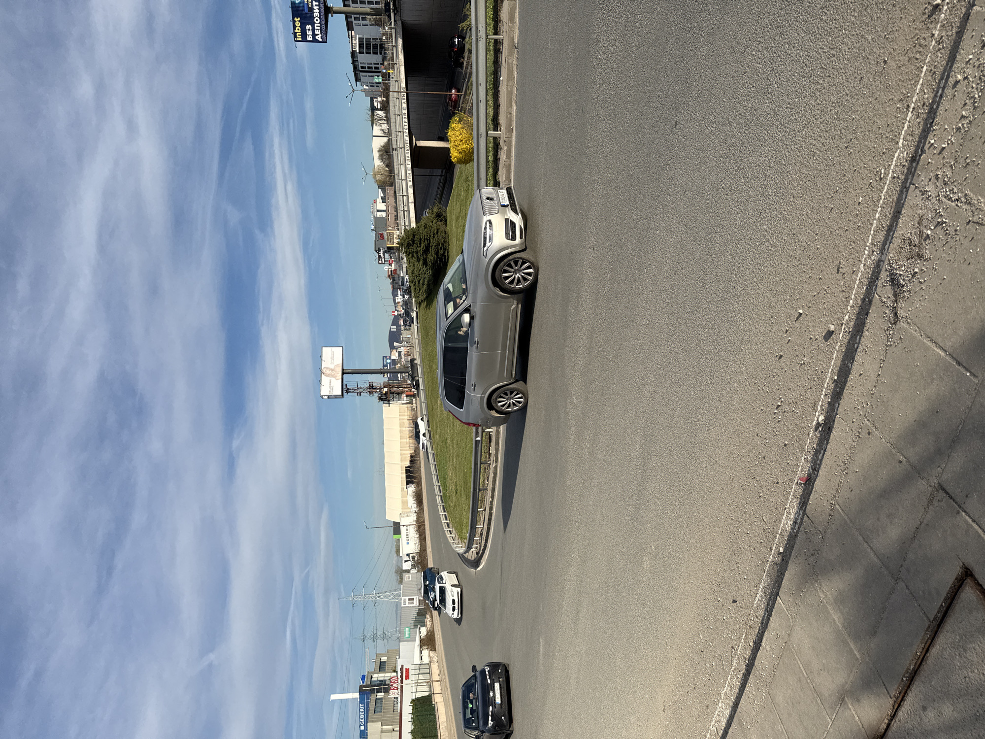 The image shows a street scene in Sofia, Bulgaria. A silver Volvo SUV is driving on an asphalt road. Other cars are also visible in the distance. On the left side of the road, there is a grassy median and various buildings. Billboards with advertisements can be seen, including one for "inbet". The sky is blue with scattered clouds. A sidewalk runs along the right side of the road. The photograph appears to have been taken during the daytime.