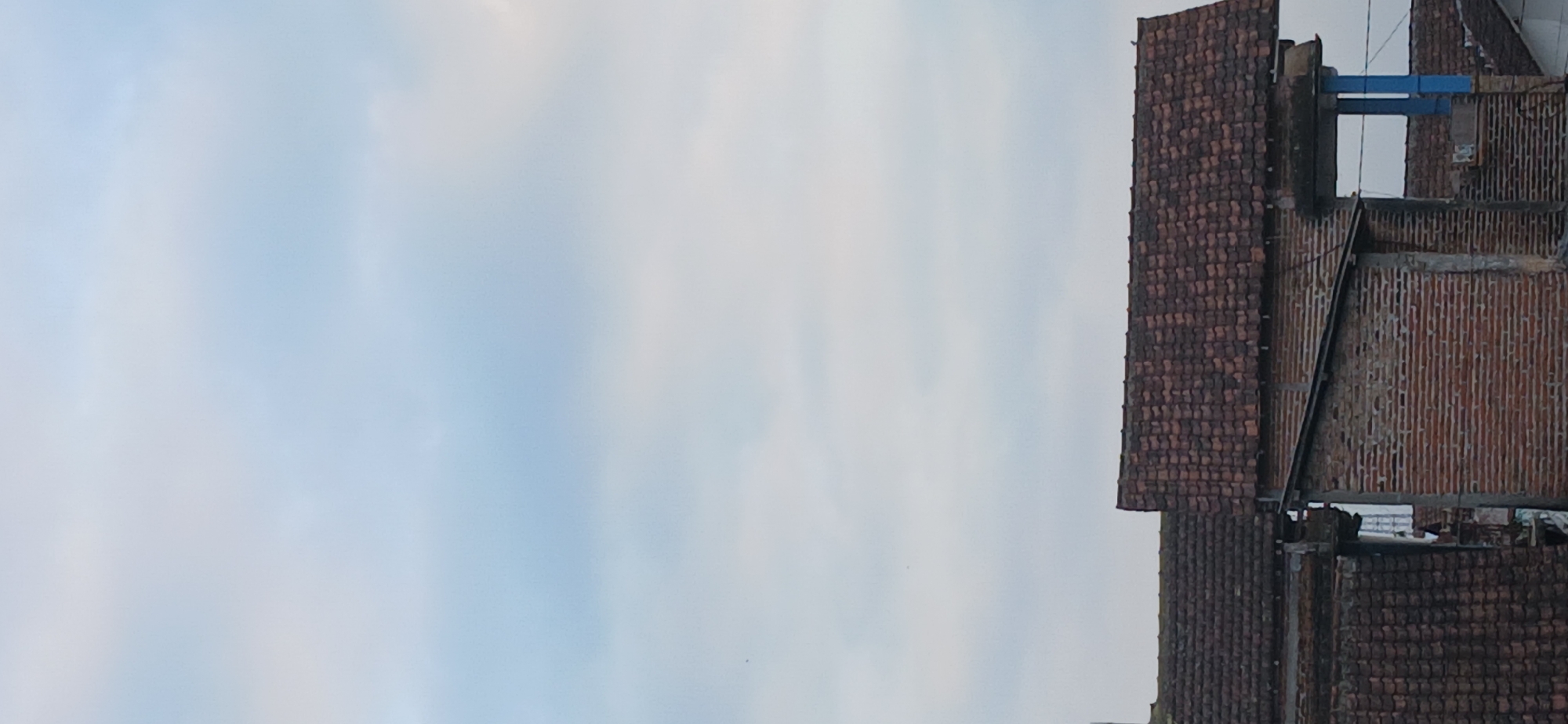 The image shows a view of several rooftops in the city of Tenggarang, Indonesia, under a cloudy sky. The rooftops are covered with traditional terracotta tiles. In the foreground, there are parts of brick buildings with visible brickwork. One building appears to have a blue support structure. The overall scene depicts a residential or urban area with older architectural elements. There are no people visible in the image. The lighting suggests it is either early morning or late afternoon, with soft, diffused light due to the cloud cover.