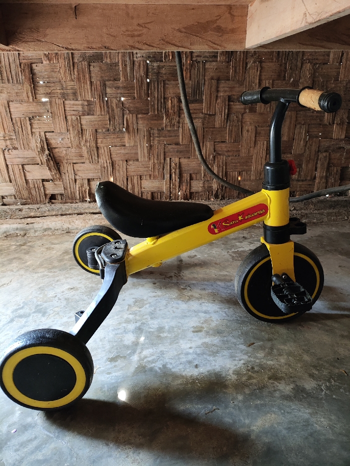 A bright yellow children's tricycle or balance bike is the central subject, positioned on a light gray concrete floor. The bike features a black seat and handlebars, with the right handlebar grip wrapped in a tan or brown material. Its wheels are black with distinctive yellow inner rims, and black pedals are visible on the front wheel. A small red plastic piece is attached to the handlebar stem. On the yellow frame, near the seat, the text "Satu Keluarga" is visible in white, accompanied by a small red logo that resembles a bird or butterfly.
The background consists of a wall made from closely woven bamboo or wicker, exhibiting a traditional construction style. Above this wall, sturdy wooden beams are exposed, indicating a rustic or sheltered structure. A dark cable or hose hangs vertically in the mid-ground. The concrete floor shows some staining and possibly damp patches, and a discernible shadow cast by the bike suggests natural daylight. The setting appears to be an indoor or semi-outdoor space, consistent with the Selagai Lingga, Indonesia location, showcasing local architectural elements. There are no people, and the tricycle is stationary.