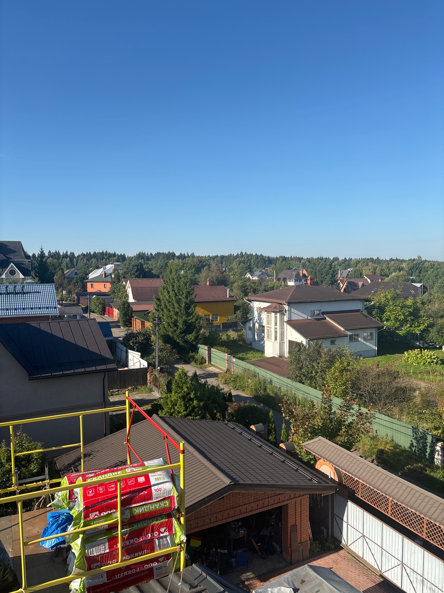 The image shows a view of a suburban area with several houses and dense green trees under a clear blue sky. In the foreground, there is a construction site with scaffolding holding stacks of building materials, including what appear to be insulation rolls wrapped in red and green packaging. The houses in the scene are of various sizes and architectural styles, some with dark-colored roofs and others with terracotta tiles. A paved road is visible winding through the neighborhood. The location is identified as Zhavoronki, Russia, suggesting a residential neighborhood on the outskirts of a city. The overall atmosphere is peaceful and suggests ongoing construction or renovation work in a rural or semi-rural setting.