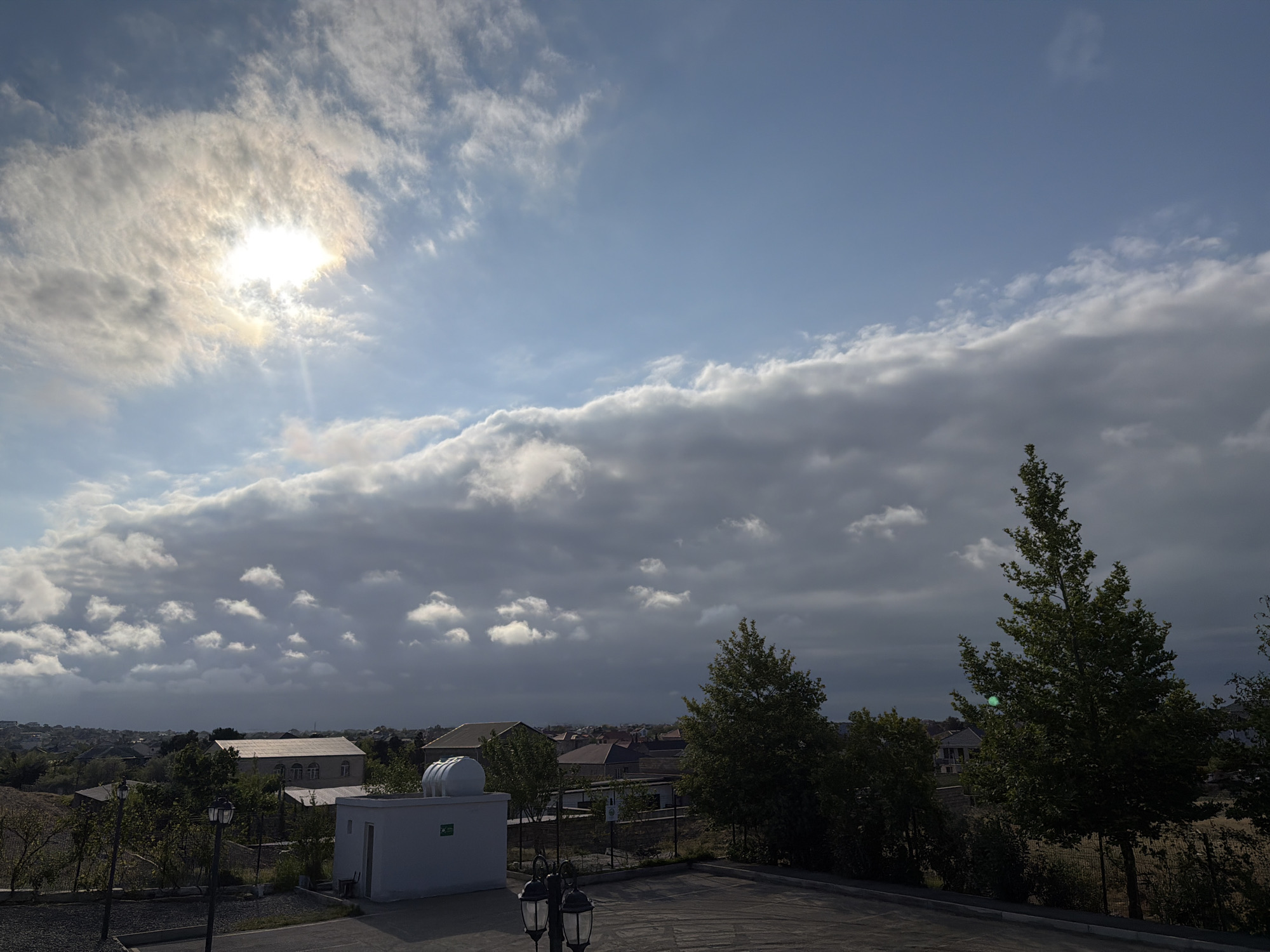 The image depicts a cityscape in Novxanı, Azerbaijan under a partly cloudy sky. The sun is visible through thin clouds in the upper left. A layer of dense, gray clouds stretches across the middle of the frame, obscuring the horizon in places and creating a contrast with the brighter sky above. In the foreground, trees and bushes line the edge of a paved area, likely a parking lot or road. A small, white, rectangular building with a curved roof feature sits to the left. Streetlights stand in the lower center, and several houses with brown roofs are partially visible amid the greenery. No people are visible.