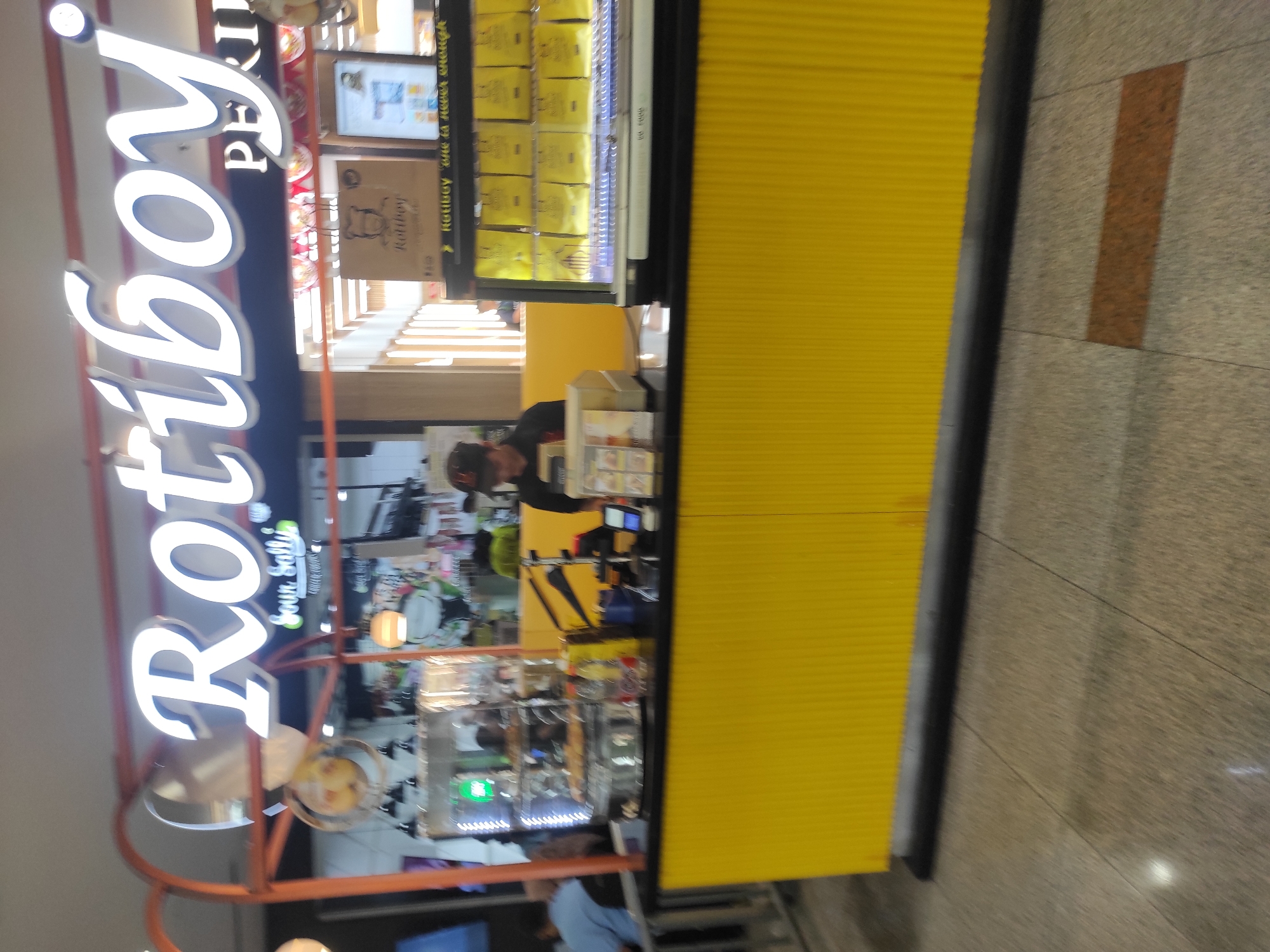 The image shows a Roti Boy store, a popular bakery chain in Indonesia, likely located in a shopping mall or public area in Surabaya. The Roti Boy logo is prominently displayed in white, backlit lettering above the store's counter. Below the main logo, a smaller sign reads "Sour Sally," suggesting a possible collaboration or adjacent store. A male employee, wearing a dark cap and black shirt, stands behind the counter, interacting with customers or preparing food. The counter itself is bright yellow with a ribbed texture, contrasting with the black countertop. To the right of the employee, a glass display case showcases rows of yellow-packaged Roti Boy products. Behind the counter, a variety of baked goods are visible in a refrigerated display case, along with various utensils and equipment. The flooring appears to be tiled with a speckled pattern. The overall atmosphere suggests a busy retail environment.