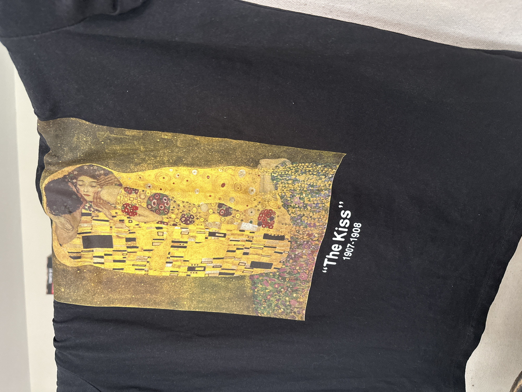 A close-up shot displays a black t-shirt featuring a print of Gustav Klimt's painting, "The Kiss" (1907-1908). The painting, characterized by its vibrant gold tones and depiction of a couple embracing, is centered on the shirt. The title and year of the painting are printed in white lettering beside the artwork. The t-shirt is photographed against a neutral backdrop. The location is not apparent and cannot be determined from the image.