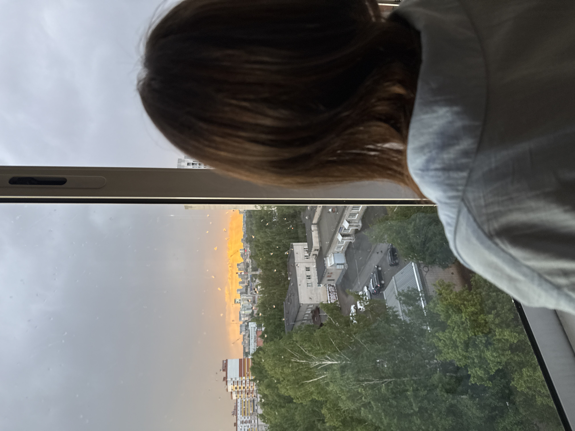 A person with short brown hair and wearing a light blue/grey button-down shirt is looking out of a window. The window has raindrops on it, and the view outside shows a cityscape with buildings, trees, and an area where there is a glow or light, seemingly from the sun or possibly a sunset or sunrise. From the position of the person, the image has been taken near the city of Izhevsk, Russia.