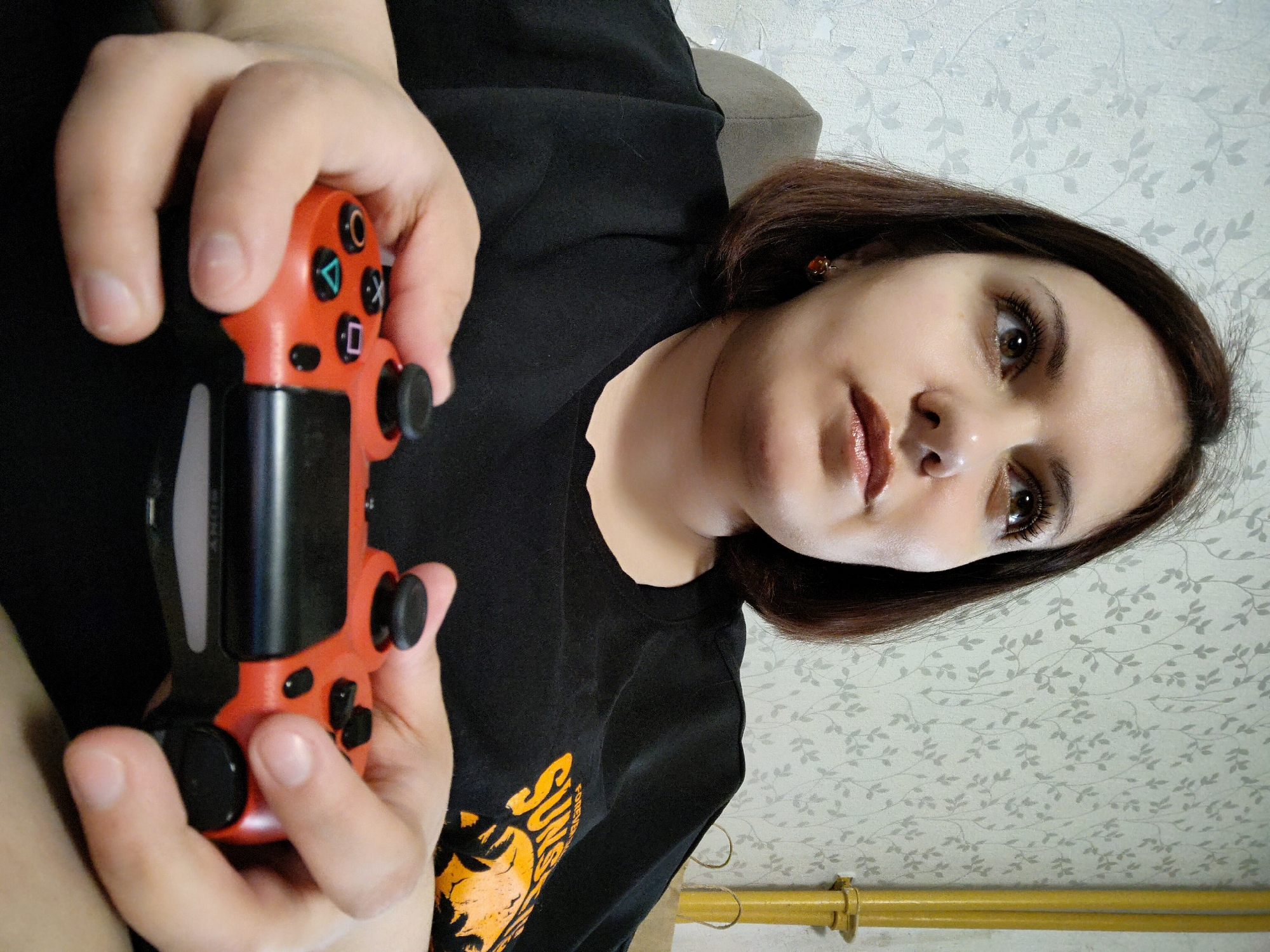 A person is holding a PlayStation DualShock 4 controller. The controller is red with black accents. The person is wearing a black t-shirt with an orange graphic. The background is a wall with a floral pattern and a yellow pipe. The image was taken in Марковский, Russia.