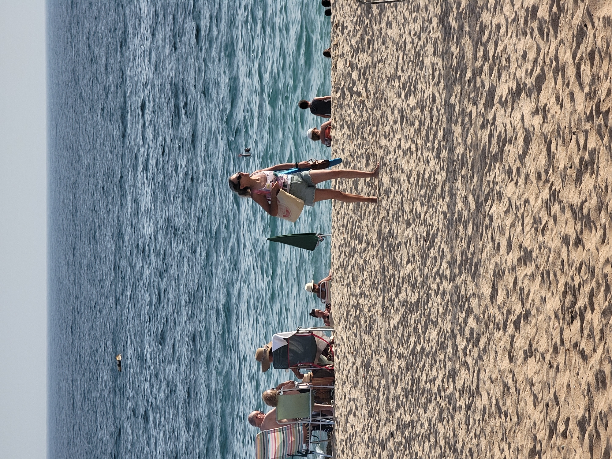 In Quarteira, Portugal, a sandy beach scene unfolds with the ocean in the background. In the foreground, a woman with blonde hair pulled back, wearing sunglasses, a patterned tank top, and light denim shorts, walks across the sand. She carries a large tote bag over her left shoulder and a blue object in her right hand. Behind her, further up the beach, a group of people are seated in beach chairs, with a green umbrella near them. In the middle distance, the blue ocean has moderate waves, and several people are visible in the water. One person is floating on their back, while another has their arm raised out of the water. Several other figures are in the water or near the shoreline. The sand is textured with ripples and shadows from the low sun.