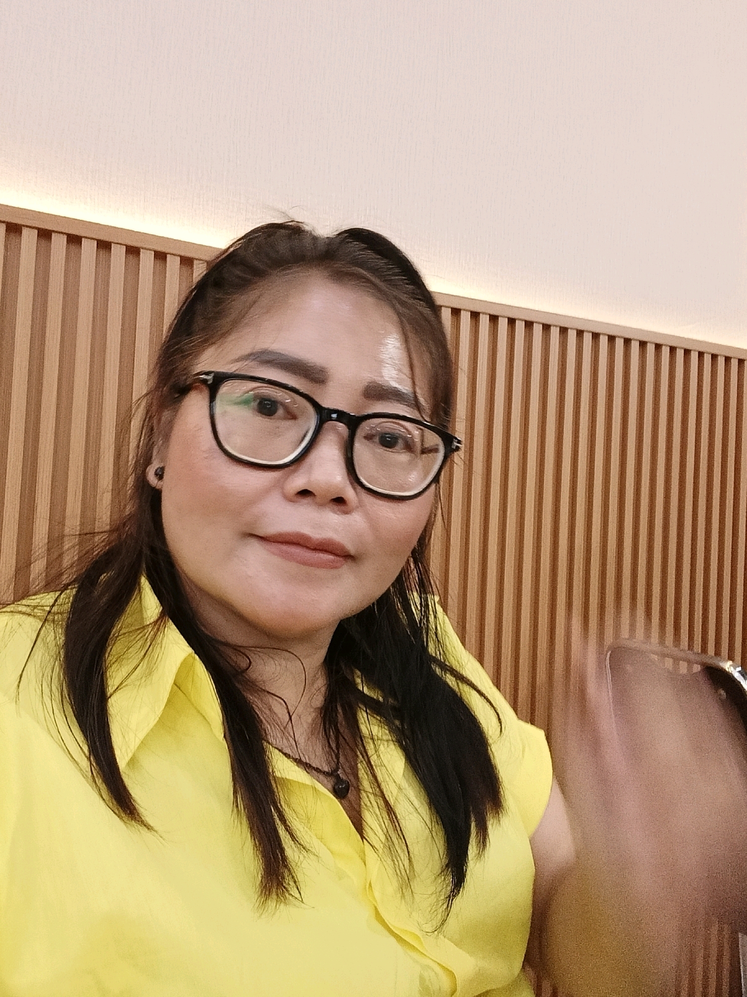 A woman with dark hair and glasses is taking a selfie indoors. She is wearing a bright yellow shirt and a dark pendant necklace. The background consists of a wall with vertical wooden slats and a soft, lit ceiling. The woman appears to be right-handed, as her right hand is blurred in the foreground, suggesting she is holding a phone to take the picture. The environment suggests a modern interior, possibly a cafe or restaurant in Jatimulya, Indonesia.