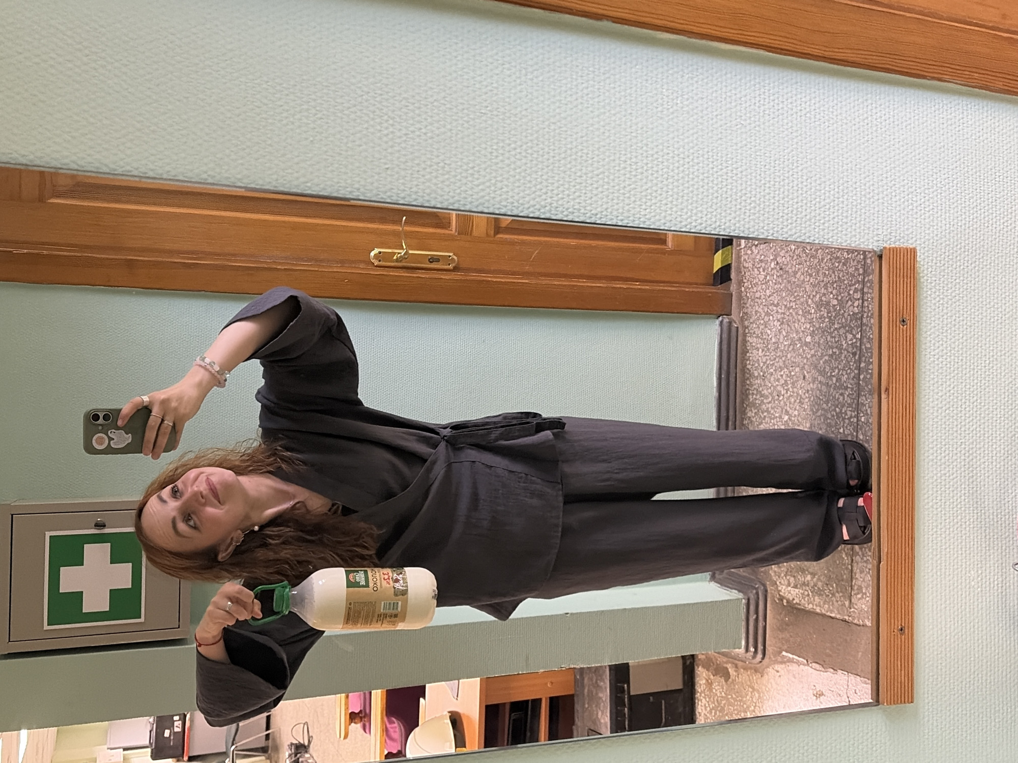 A woman with shoulder-length brown hair, wearing a dark grey wrap top and matching pants, stands in front of a mirror and takes a selfie with her smartphone. She holds a clear plastic bottle of milk in her right hand. The bottle has a green cap and a label with Russian text. The woman is also wearing black sandals with a red accent. Behind her, reflected in the mirror, is a first-aid kit mounted on a pale green wall. To the right of the mirror, a wooden door with a brass handle is visible. The scene appears to be indoors, possibly in a locker room or office, given the presence of the first-aid kit and the general utilitarian aesthetic of the environment. The flooring is a speckled grey and white tile.