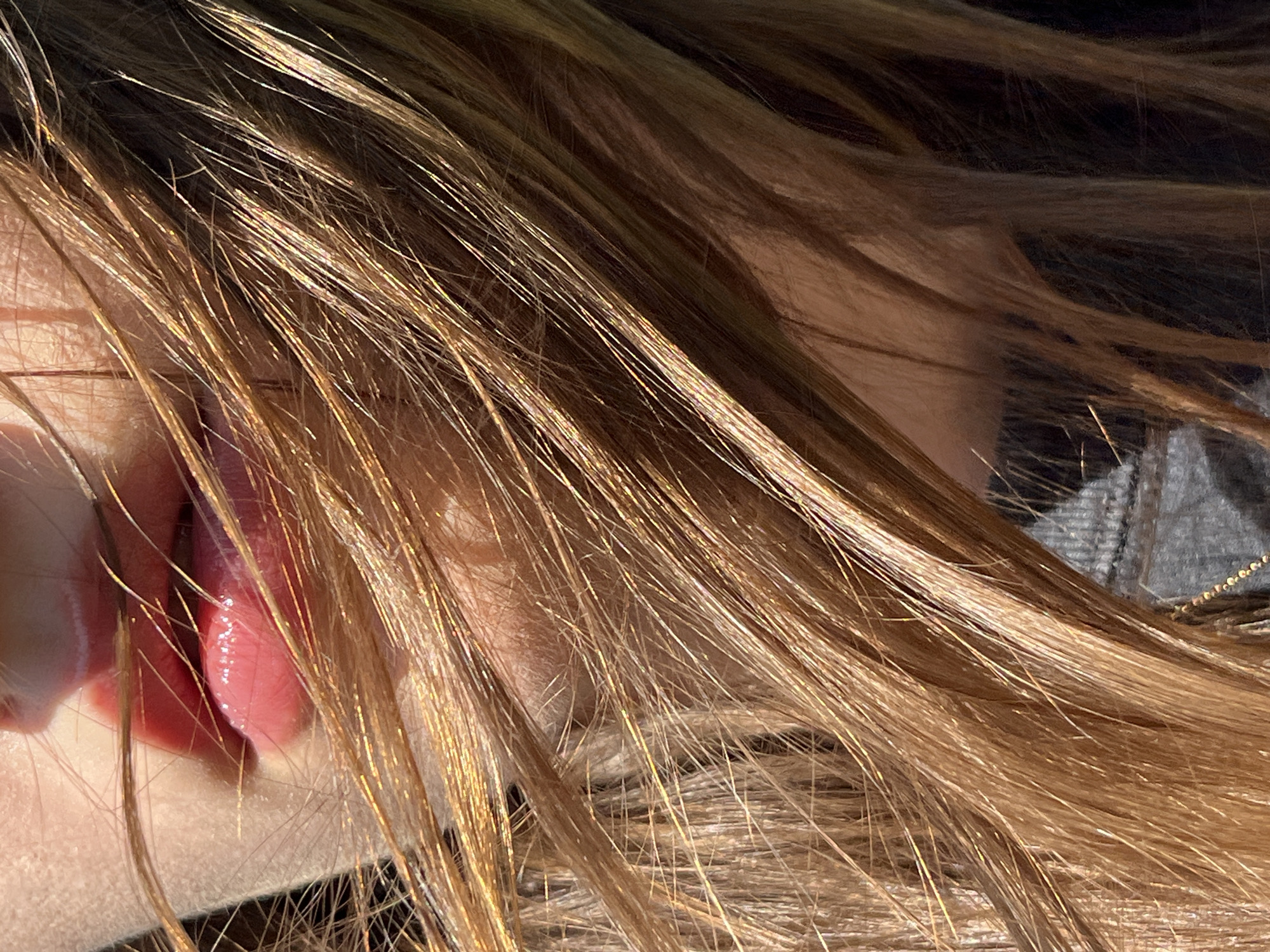 This is a close-up, top-down shot of a person's face, with their hair prominently featured. The hair is a light brown with golden highlights, catching the sunlight and creating a shimmering effect. The individual's face is partially obscured by their hair, but their mouth is visible. The lips are pink and appear glossy, with the lower lip slightly parted and the tongue visible, appearing wet and pink. The person's skin is fair. The background is dark and out of focus, with some suggestion of clothing, possibly a textured fabric like denim or tweed, and a glimpse of a chain or necklace. The lighting suggests it is daytime, with the sun casting strong shadows and illuminating the hair. The overall impression is intimate and sensual. There is no visible text.