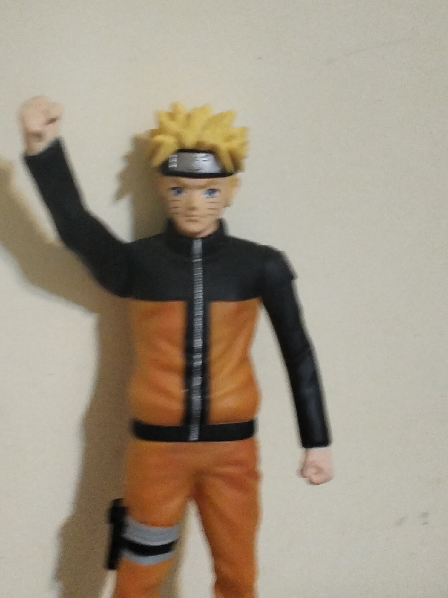 A figurine of the anime character Naruto Uzumaki is displayed against a plain, light-colored wall. The figurine depicts Naruto from the waist up, with his left arm raised in a fist. He is wearing his signature orange and black outfit, including a black headband with a silver plate. The background is a plain, textured wall with a subtle shadow cast by the figurine on the left side. There is no indication of the location being in Baku, Azerbaijan; it appears to be an indoor setting.
