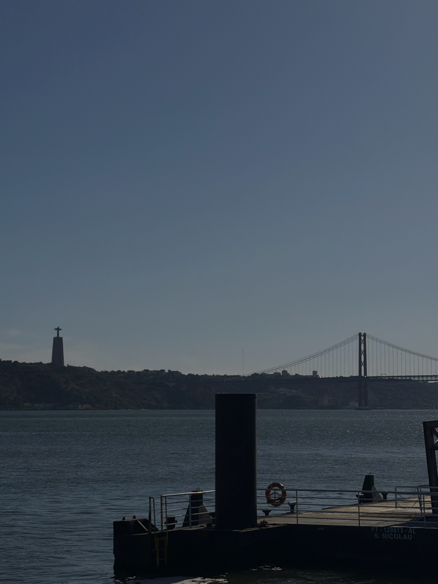 The image depicts a wide shot of the Tagus River in Lisbon, Portugal, under a clear blue sky. In the distance, silhouetted against the horizon, is the 25 de Abril Bridge, a prominent suspension bridge spanning the river. To the left of the bridge, on a hilltop, stands the Cristo Rei (Christ the King) statue, also silhouetted. In the foreground, a dark, flat-topped barge or floating platform is visible. On the platform, a thick, dark cylindrical bollard stands on the left, and a life preserver ring with an orange hue is attached to a railing. Further to the right on the platform, railings and other structures are present. Text is visible on the side of the platform, reading "PT-129613-AL S. NICOLAU". The water of the river is a deep blue-grey, with gentle ripples. The scene suggests a calm day on the river, with the iconic landmarks of Lisbon in the background and maritime infrastructure in the foreground.
