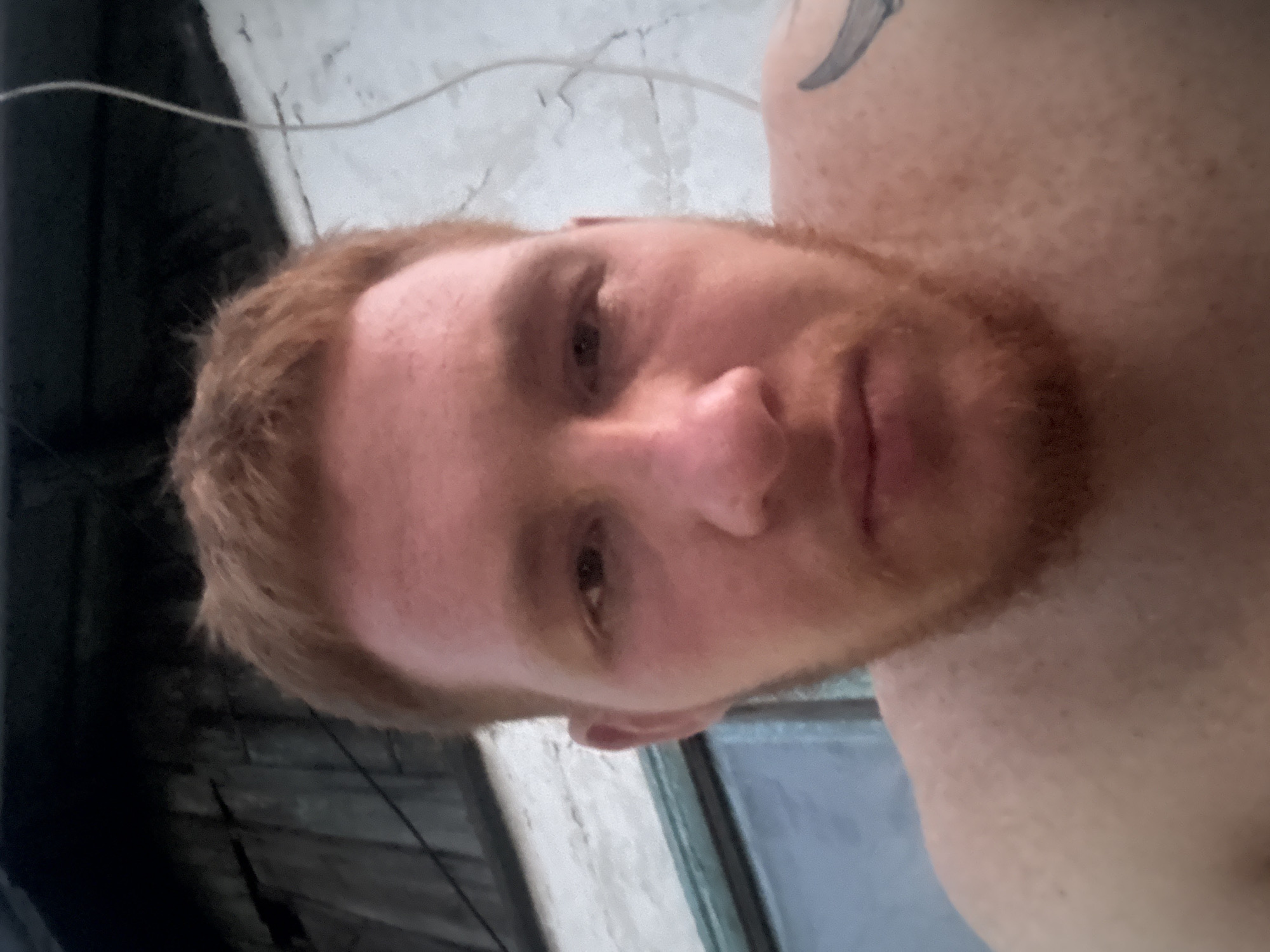A close-up, waist-up, outdoor selfie taken by a fair-skinned man with short, reddish-blonde hair and a beard of the same color. The man's eyes are brown, and his skin is flushed. He is looking directly at the camera, with his mouth slightly open. His left shoulder is visible, and a portion of a dark tattoo is present on his upper left shoulder. The background is out of focus, but appears to be a weathered wooden structure on the left and a textured white wall on the right, with a thin, light-colored wire extending from the top right corner diagonally downwards. The lighting suggests it may be dusk or dawn. The location is Unknown, Unknown.