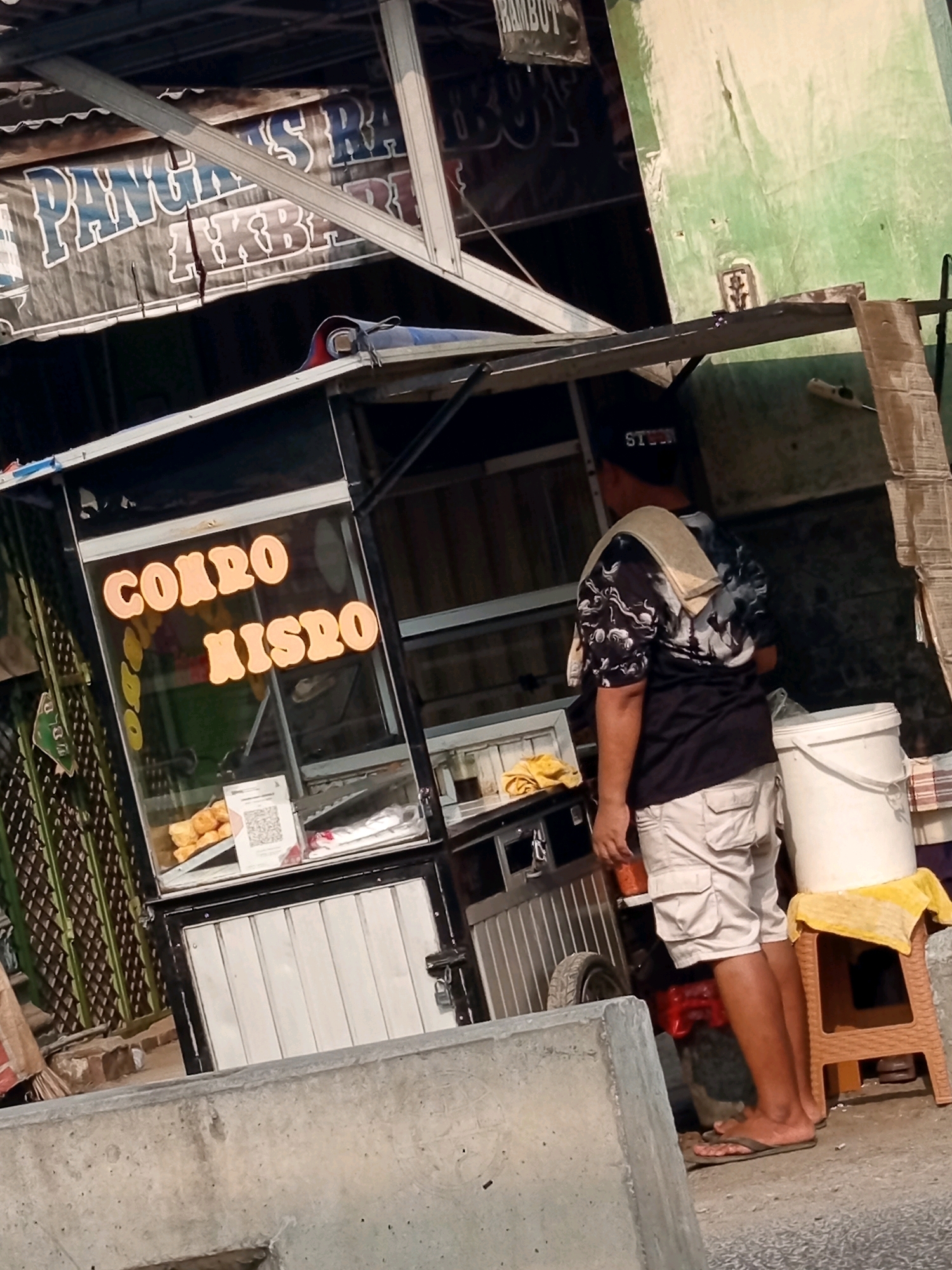 The image depicts a street food stall in Karawang, Indonesia. A man wearing a black t-shirt with a white graphic pattern, light-colored cargo shorts, and a dark baseball cap is standing behind the stall. He has a beige towel draped over his left shoulder. The stall has a glass display case showing food items, a QR code for payment, and a white bucket on a small brown stool. Above the stall, a banner with blue and white text is visible, partially obscured by metal framing. Another banner with black text is mounted higher up. The stall is situated outdoors, with a concrete barrier in the foreground and a green wall in the background. The overall scene suggests a typical urban street food vending setup.