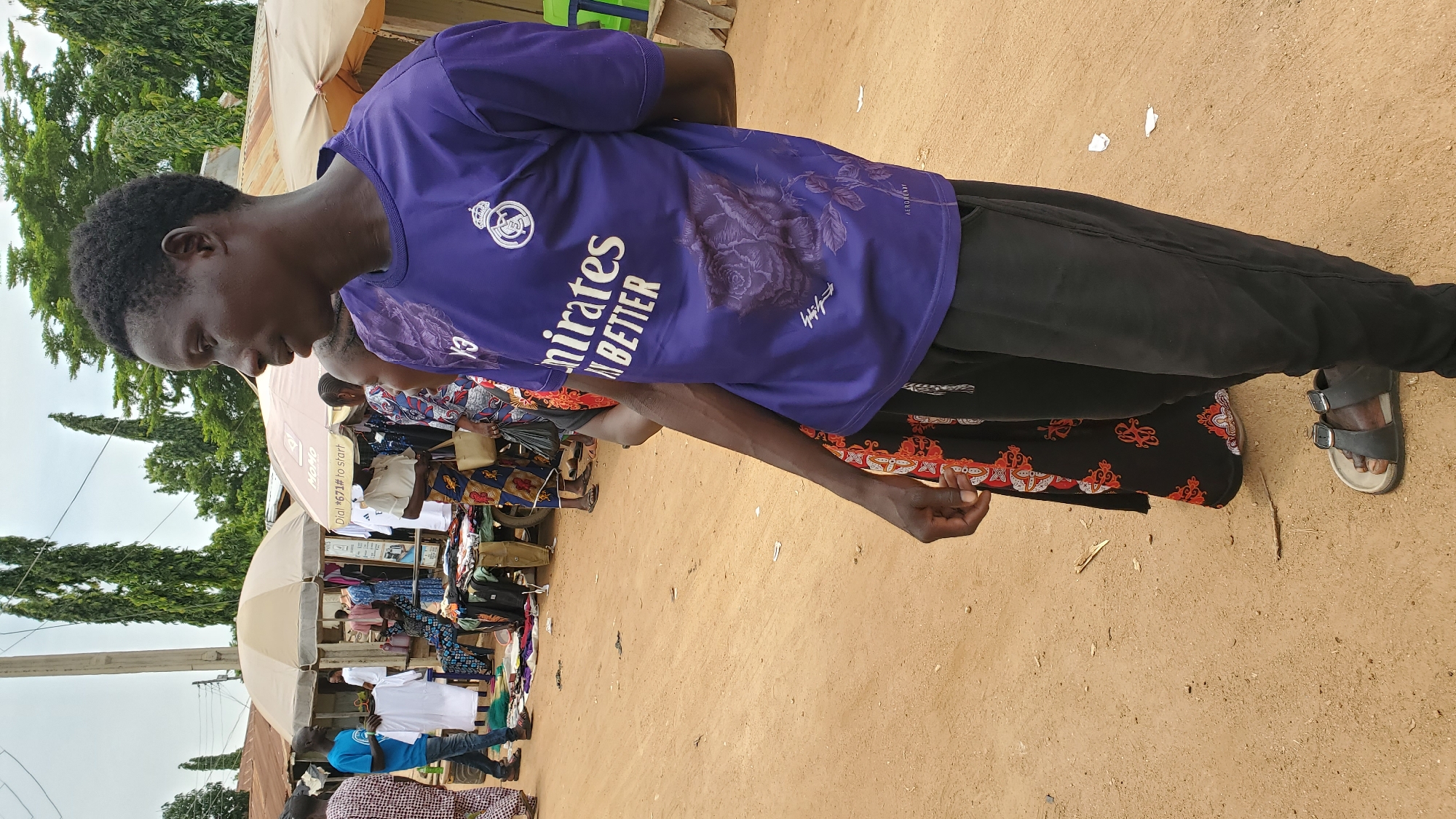 The image depicts a street market scene in Jalingo, Nigeria. In the foreground, a Black man wearing a purple t-shirt with the "Emirates" logo and a floral design, and black pants, is standing with his back slightly turned to the camera. He is wearing dark sandals. Beside him, to his left, a person wearing a black skirt with a red and white pattern is visible. The background shows various market stalls with umbrellas and awnings. People are seen browsing and interacting at the stalls, with clothing and goods displayed for sale. The ground is unpaved and dusty, consistent with a rural or semi-urban environment. Tall trees are visible in the distance. The overall atmosphere suggests a typical day at a local market.