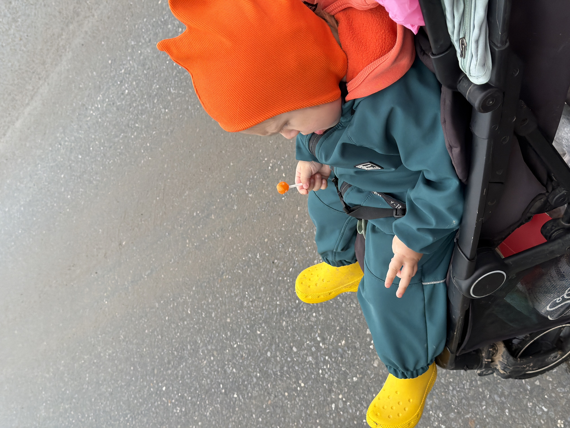 A child wearing an orange hat and yellow rain boots is seated in a stroller on a paved surface. The child is holding a lollipop. The stroller is black and has a light blue bag hanging from it. The paved surface appears to be a sidewalk or street. The location is Chaikovsky, Russia.