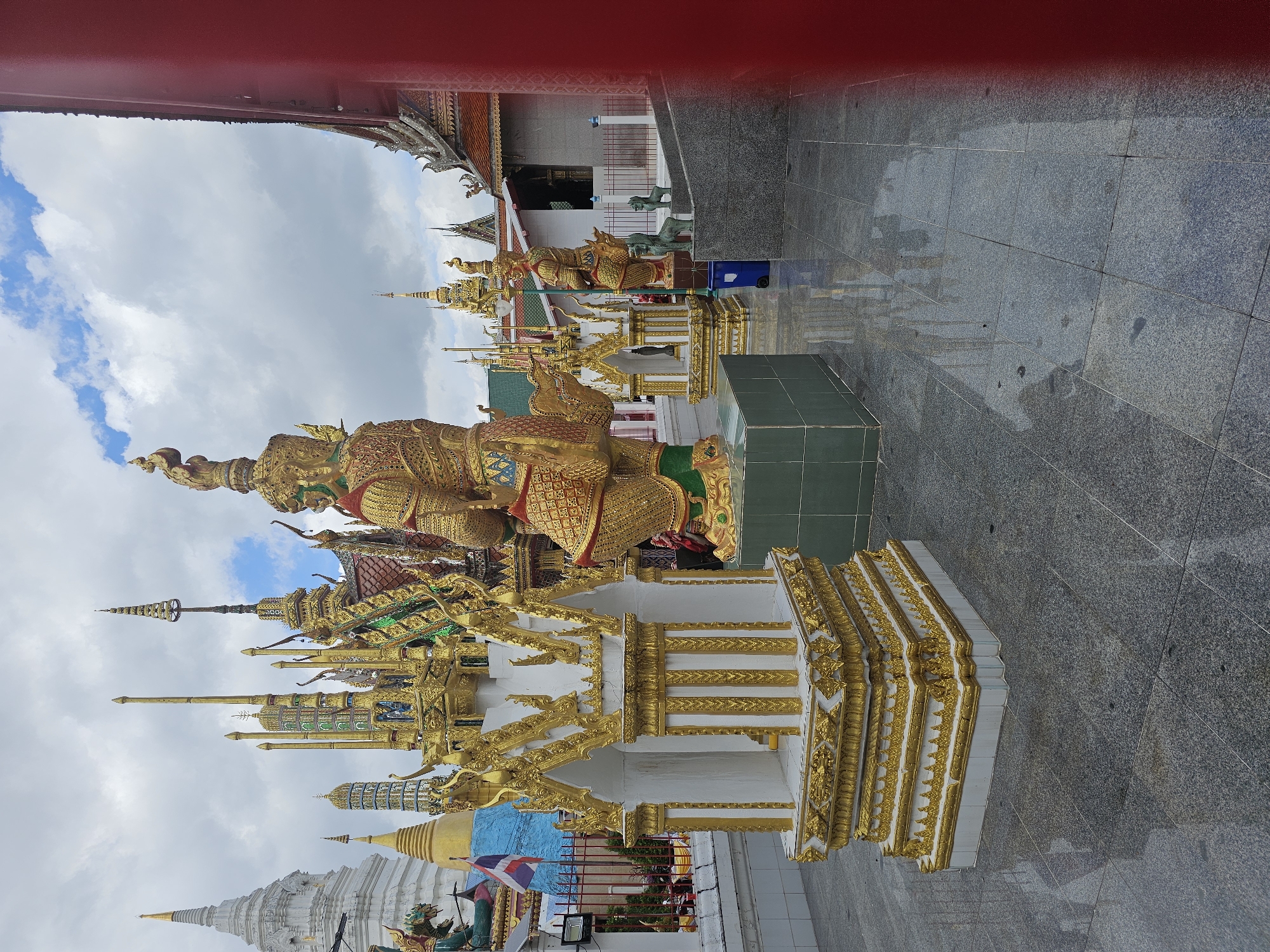The image depicts a courtyard with ornate statues and temple architecture, likely in Bangkok, Thailand. In the foreground, a small, white structure with gold embellishments stands on a patterned stone floor. To the right, a large, golden statue of a warrior or deity is visible, adorned with intricate details. In the background, several multi-tiered structures with golden spires and colorful roofs rise against a cloudy sky. A white stupa with a golden spire is also prominent. The ground is wet, reflecting the surrounding structures. The scene appears to be a religious or cultural site, possibly a Buddhist temple.