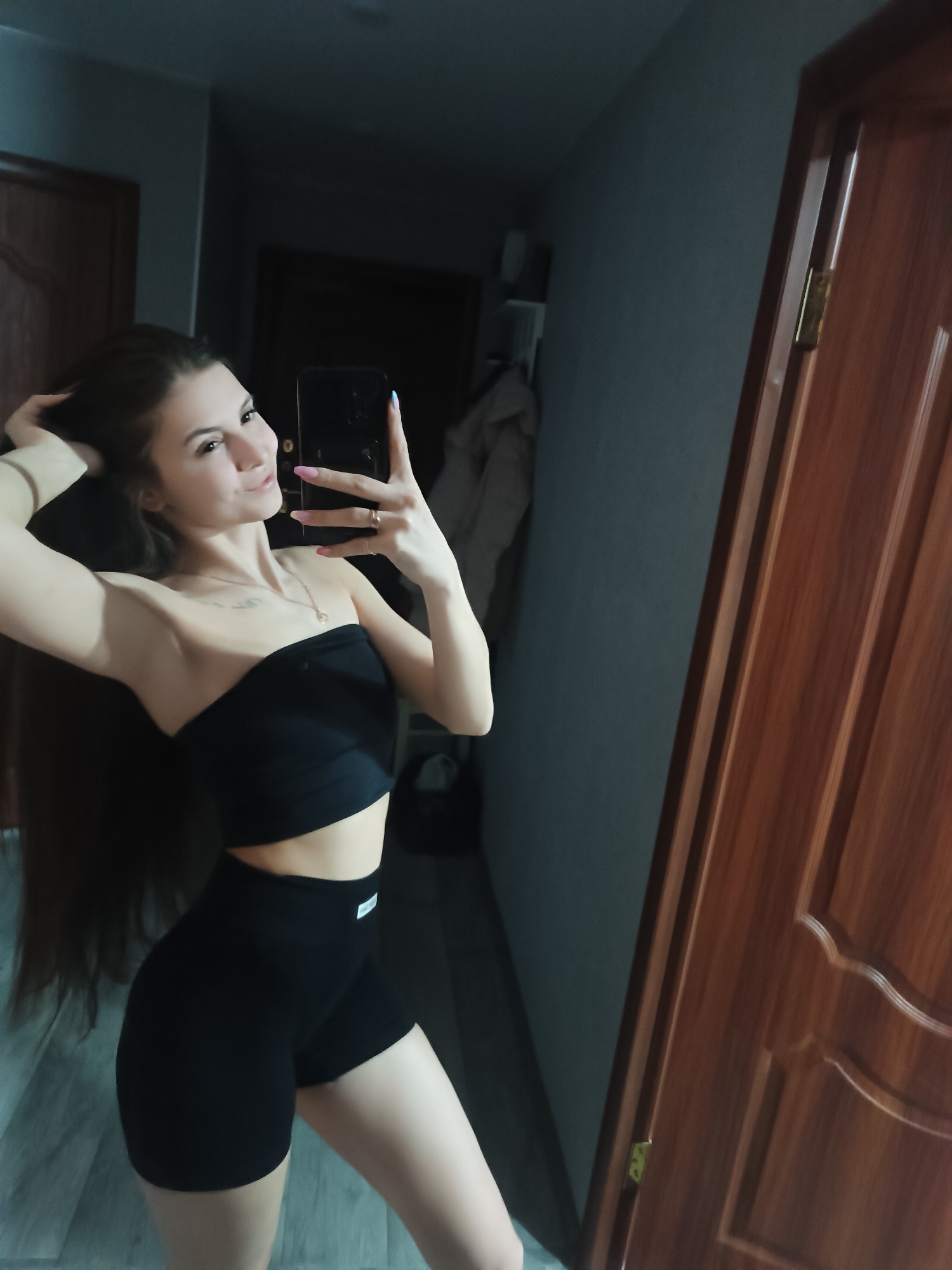 A woman is taking a selfie in what appears to be an indoor environment. She is wearing a black strapless crop top and black shorts. Her hair is long and dark. She's holding a smartphone in her right hand, using it to capture the image in a mirror. The background includes a wooden door and what seems to be a hallway. The location, according to the prompt, is Chaikovsky, Russia.