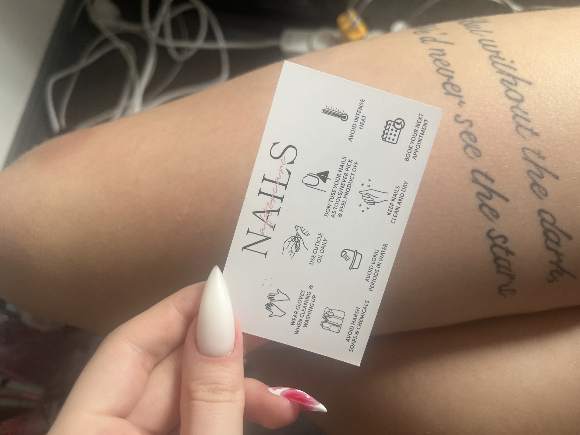 A person is holding a white business card for a nail salon with the word "NAILS" printed on it. The card lists nail care instructions such as "Avoid Intense Heat," "Don't use your nails as tools/Never pick & peel product off," and "Keep nails clean and dry". The person has long, white stiletto nails. A tattoo is visible on the person's arm. The location in Varna, Bulgaria is not possible to ascertain from the image.