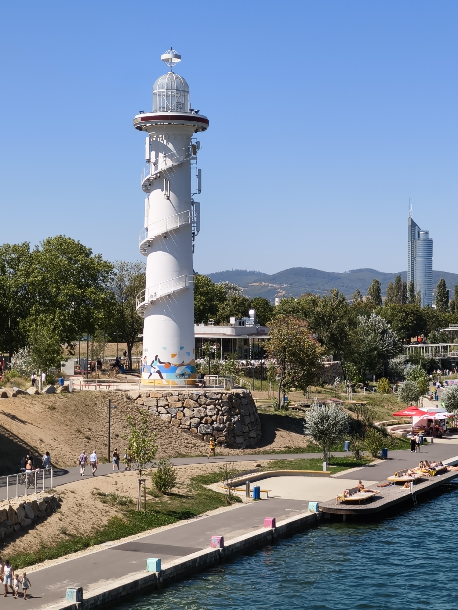 The image depicts a sunny day at the Danube River in Vienna, Austria. In the foreground, a modern white lighthouse with a spiral staircase stands prominently. The lighthouse is decorated with a colorful mural depicting people playing sports. The surrounding area features a waterfront promenade, lush green trees, and a modern building complex in the background, including a distinctive skyscraper. People are seen walking along the promenade, cycling, and relaxing on loungers on a wooden pier extending into the water. The blue water of the Danube river reflects the clear blue sky. The scene suggests a recreational area with various amenities for visitors to enjoy outdoor activities and leisure.
