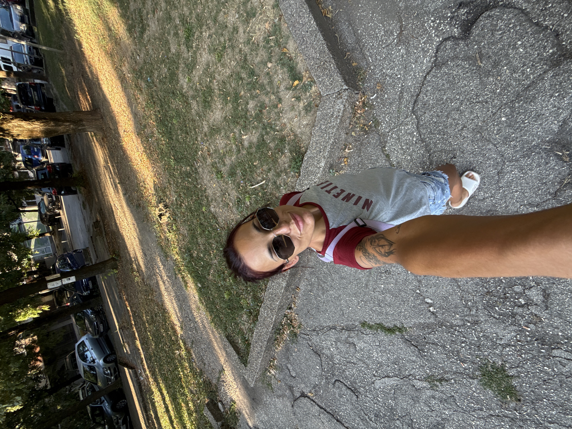 A woman is taking a selfie outdoors in the city of Pleven, Bulgaria. She is wearing a gray t-shirt with a burgundy neckline and sleeves, denim shorts, and white sandals. She has on large, dark sunglasses and her dark hair is pulled back. She has a tattoo on her left arm. The background shows a sidewalk, a grassy area, and parked cars. The image appears to have been taken on a sunny day.