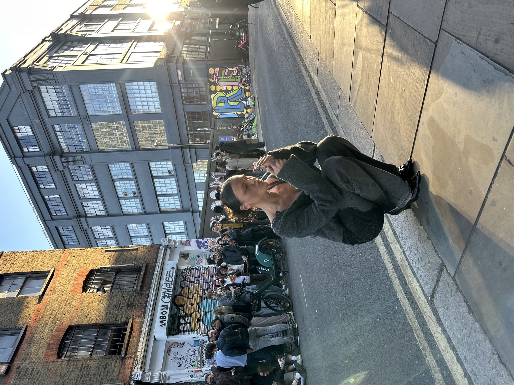 The image captures a vibrant urban street scene in London, likely during late afternoon or early morning, characterized by the low sun casting bright highlights and long shadows across the pavement and buildings.
In the immediate foreground, a person with dark hair, wearing a dark grey top and trousers, is seated on the grey tiled pavement in an unusual, contorted pose. They appear to be smoking a cigarette or similar object, holding a white stick to their mouth with a wisp of smoke visible, and are looking upwards.
To the left, a gathering of people lines the sidewalk in front of a brick building. Many individuals in the crowd are observing something out of frame or socializing. A green "Forest" rental bicycle is visible among them. Further down the street, several motorbikes or scooters are parked.
The background is dominated by a multi-story, grey industrial-style building featuring numerous large, gridded windows. Its lower section is adorned with colorful street art, including a prominent graffiti piece with letters that resemble "RI" or "RN" in blue, yellow, and pink, interspersed with smiley faces. On the closer brick building, black graffiti text reads "1980 M CALISE" (partially obscured). The street features asphalt with a double white line, bordered by the grey tiled pavement. The sky is bright and clear, with a visible sunburst from the low sun.