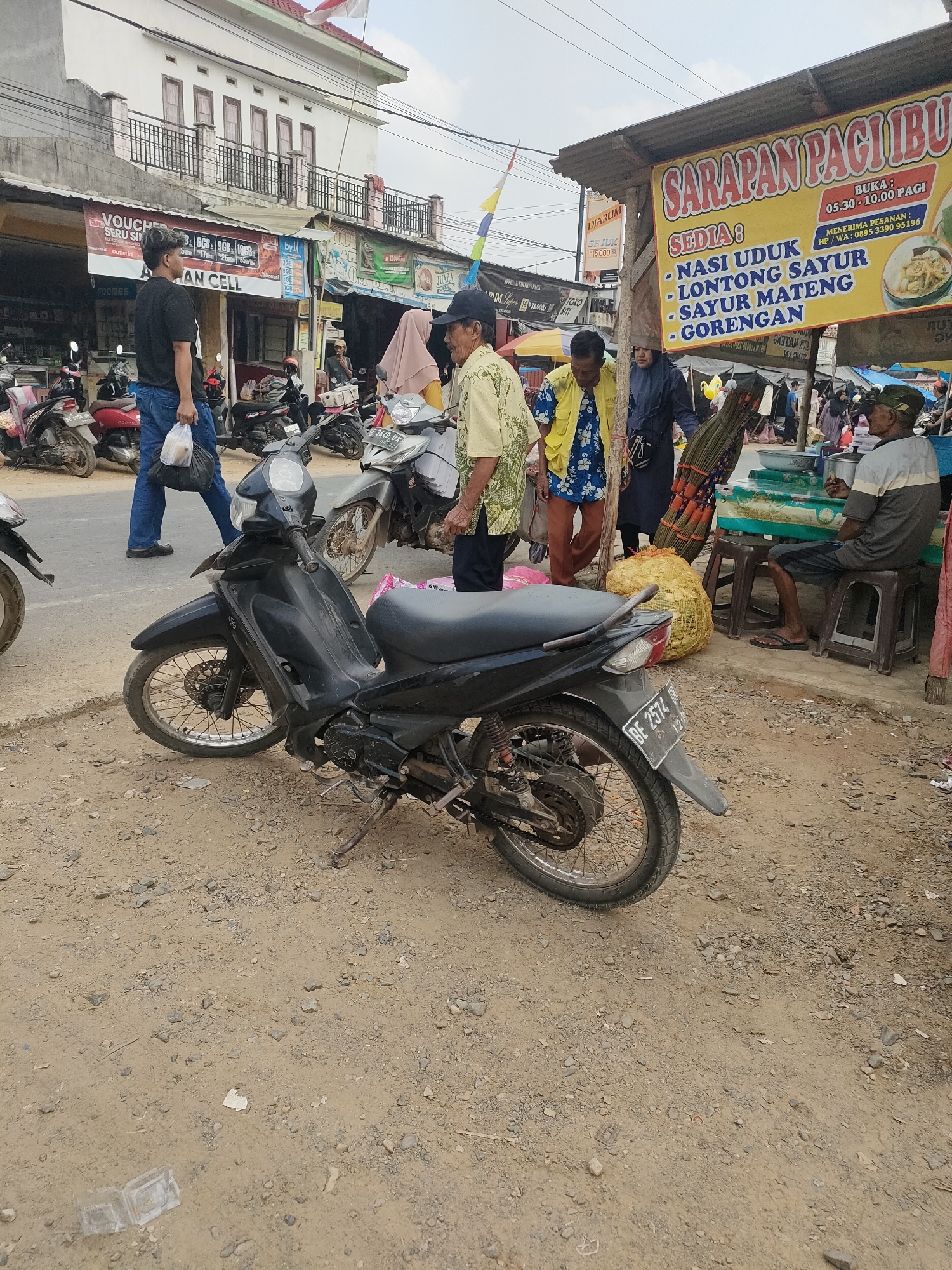 The image depicts an outdoor market scene in Sukoyoso, Indonesia, on a cloudy day. In the foreground, a black motorcycle is parked on a dirt and gravel surface. The motorcycle's license plate reads "BE 2574". To the left of the motorcycle, a man wearing a black t-shirt and blue jeans walks away from the camera, carrying a plastic bag in his right hand.
Behind the motorcycle, a yellow and white banner, advertising "SARAPAN PAGI IBU", is prominently displayed, indicating breakfast offerings. The banner lists menu items such as "NASI UDUK", "LONTONG SAYUR", "SAYUR MATENG", and "GORENGAN". Below the banner, a man in a yellow shirt and a woman in a blue headscarf stand near a stall. The stall has green countertops and appears to be selling food or goods. To the right, an elderly man wearing a cap and a gray and white striped shirt sits on a plastic stool, facing a table.
In the background, several other motorcycles are parked. Buildings with white facades and balconies line the street. A red and white Indonesian flag flies from a building on the left. The overall environment suggests a bustling local marketplace with food vendors and shoppers.