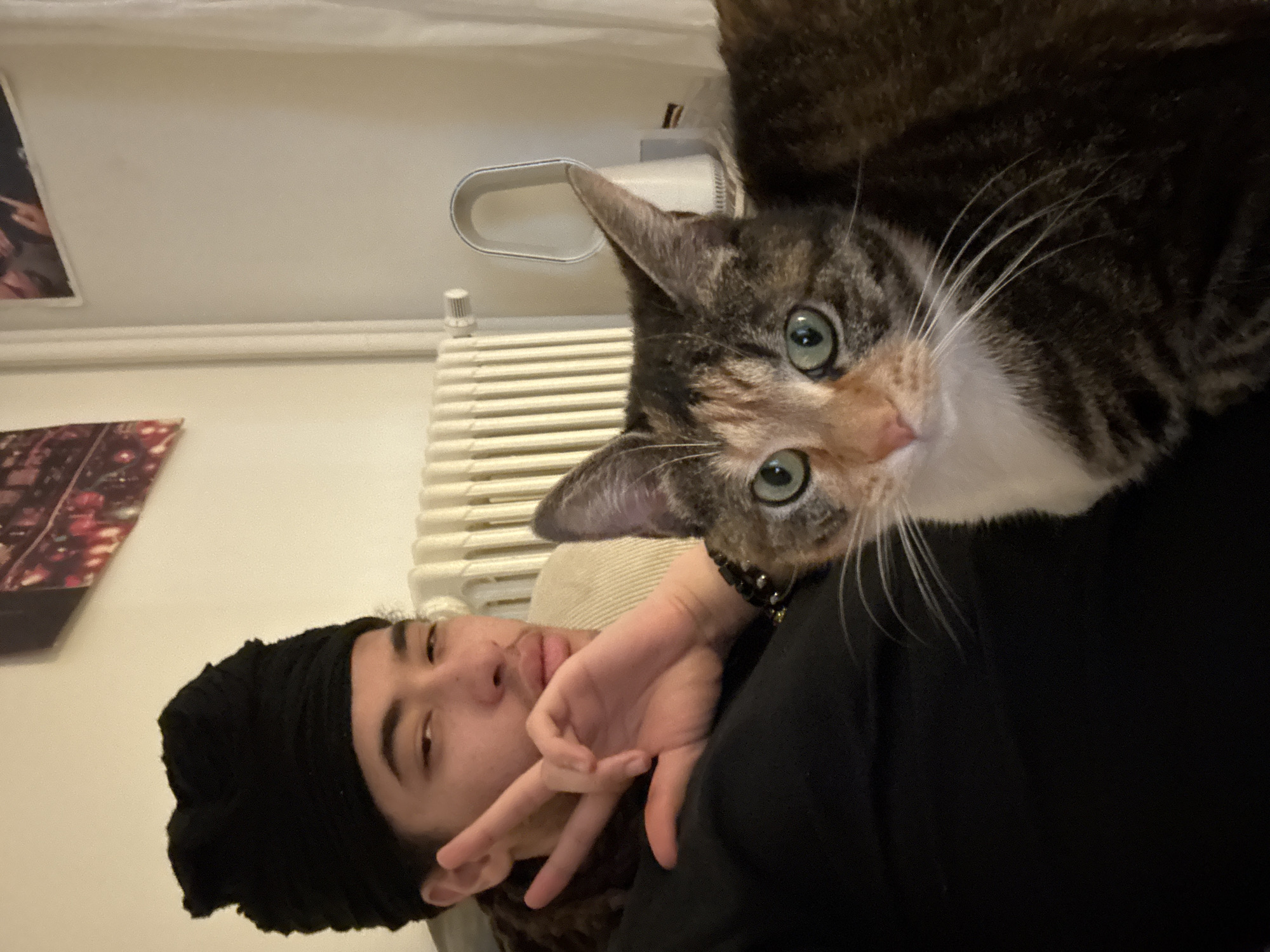 An indoor scene, presumed to be located in Paris, France, features one person and one cat. A young adult individual with a black fabric headwrap and a dark long-sleeved top is positioned on the left, visible from the chest up. Their right hand is raised, forming a "peace" sign, and a dark beaded bracelet is present on their wrist. They have a mustache and light stubble, looking directly at the camera. To the right of the individual, a domestic shorthair cat with a tortoiseshell and white coat and green eyes sits or lies, also facing the camera. The background consists of a light-colored wall with a white ribbed radiator partially visible on the left. Two framed artworks are partially visible on the wall above the radiator, one depicting a night cityscape. A white bladeless fan is visible on a surface to the right, next to a sheer white curtain. A beige cushion is visible behind the person's upper body.