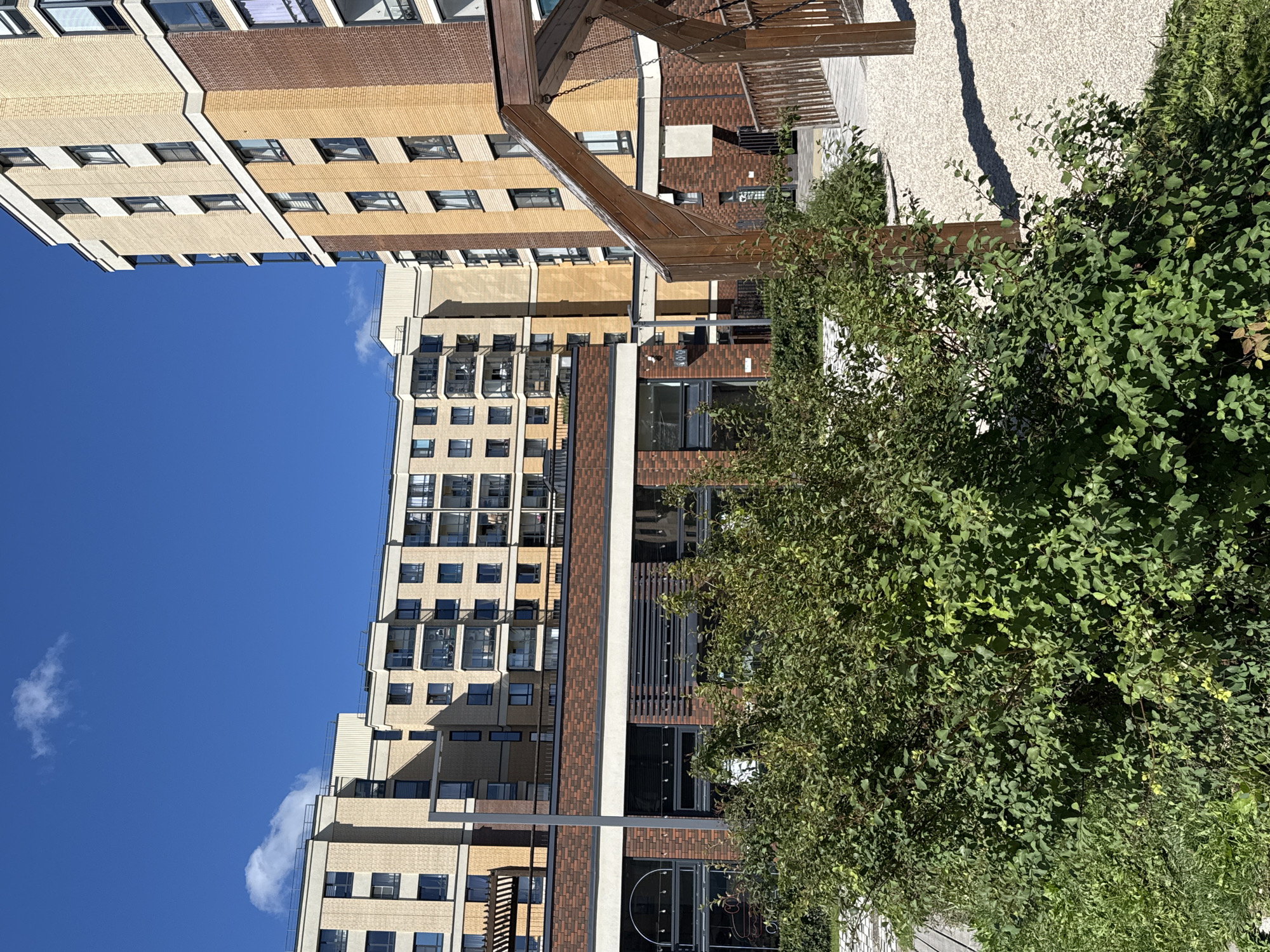 The image depicts a modern residential complex in Novoselye, Russia, under a clear blue sky with scattered white clouds. The architecture features multi-story buildings constructed with a combination of light beige brick and darker brick accents. The buildings have numerous windows and balconies. In the foreground, lush green bushes and some patchy grass are visible. A wooden play structure, possibly part of a playground, is present on the right side of the frame. The ground in the playground area is covered with light-colored gravel. The overall scene suggests a well-maintained urban environment.