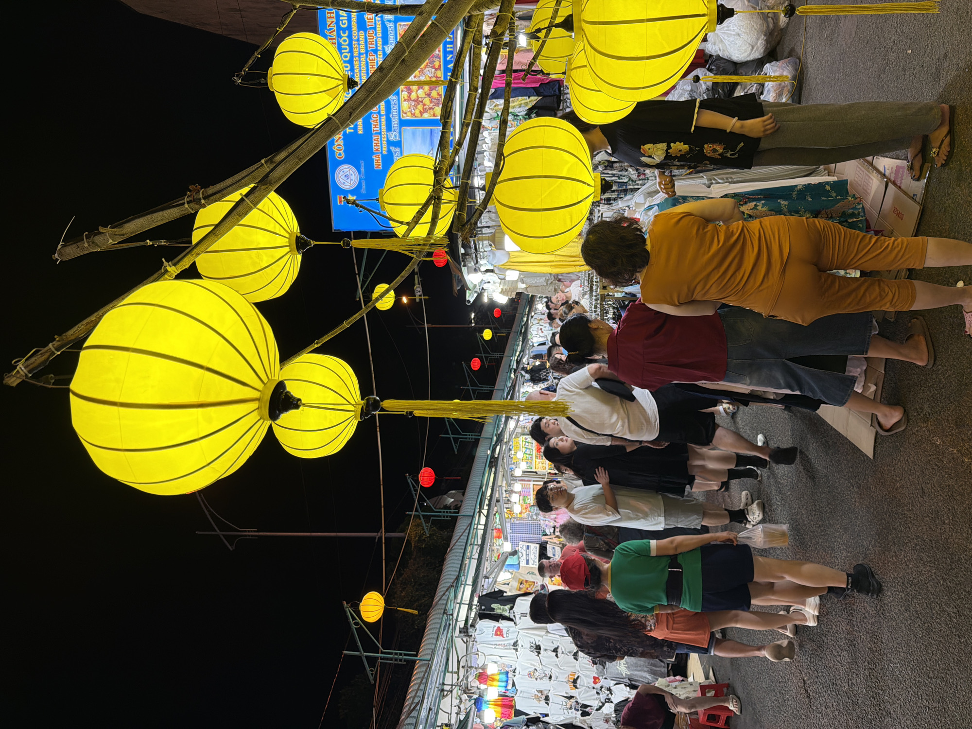 This is a photograph taken at night, likely in Nha Trang, Vietnam, depicting a bustling night market. The scene is illuminated by numerous bright yellow lanterns, which are the dominant objects in the foreground, hanging from bamboo poles. These lanterns are spherical and feature a ribbed design. In the background, a market stall is visible, with racks of clothing, predominantly t-shirts with various prints.
The photograph features several people, appearing to be a mix of locals and tourists, browsing the market. Their clothing suggests a warm climate. The people are casually dressed, and their postures indicate they are walking and looking at the goods on display. The overall atmosphere seems lively and perhaps a bit crowded, typical of a night market environment.
Visible text in the image, though partially obscured and rotated, includes Vietnamese characters that translate to "National Brand," "Professional Bird," and "Company." This text, along with the style of the lanterns and the general setting, strongly suggests a Vietnamese location. The time of day is clearly night, indicated by the dark sky and the artificial lighting from the lanterns and stall. There are no obvious cues about the weather, but the presence of people outdoors suggests it is likely fair.