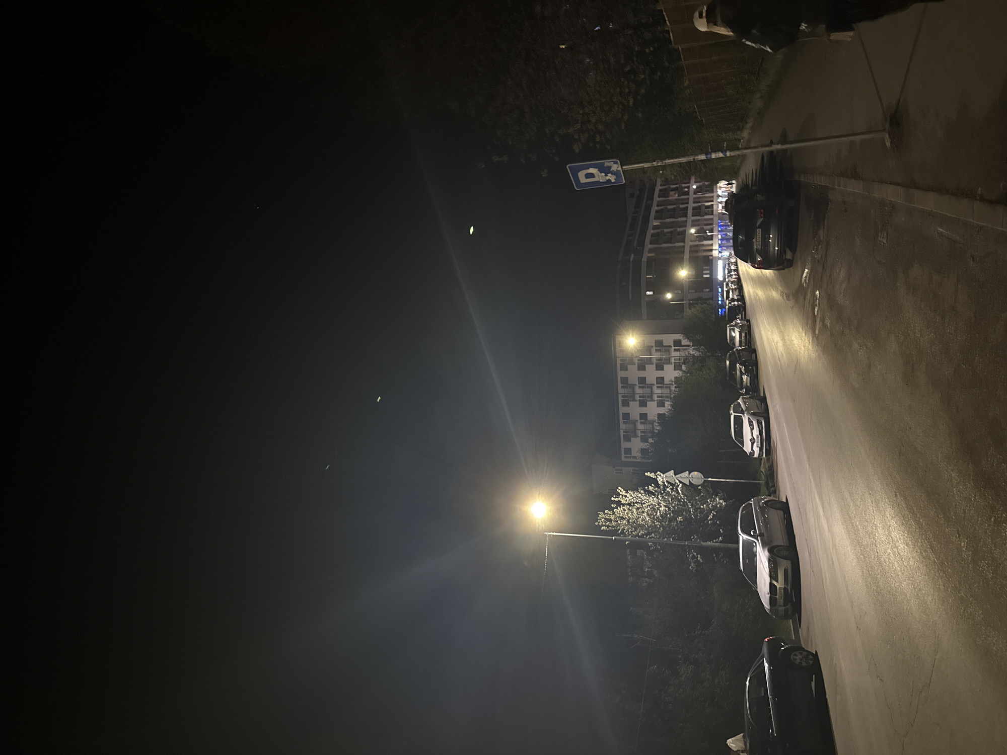 The image shows a nighttime street scene in Sofia, Bulgaria. The photo is taken from a standing height looking down the street. The road is lined with parked cars on one side and a sidewalk. A street light illuminates the road. A building can be seen in the background. There are no visible people in the immediate foreground, though one is partially visible at the upper edge of the image, possibly walking. There are some trees along the street. A parking sign is visible in the background. There are two small bright spots in the sky, one near the parking sign and one near the street lamp.